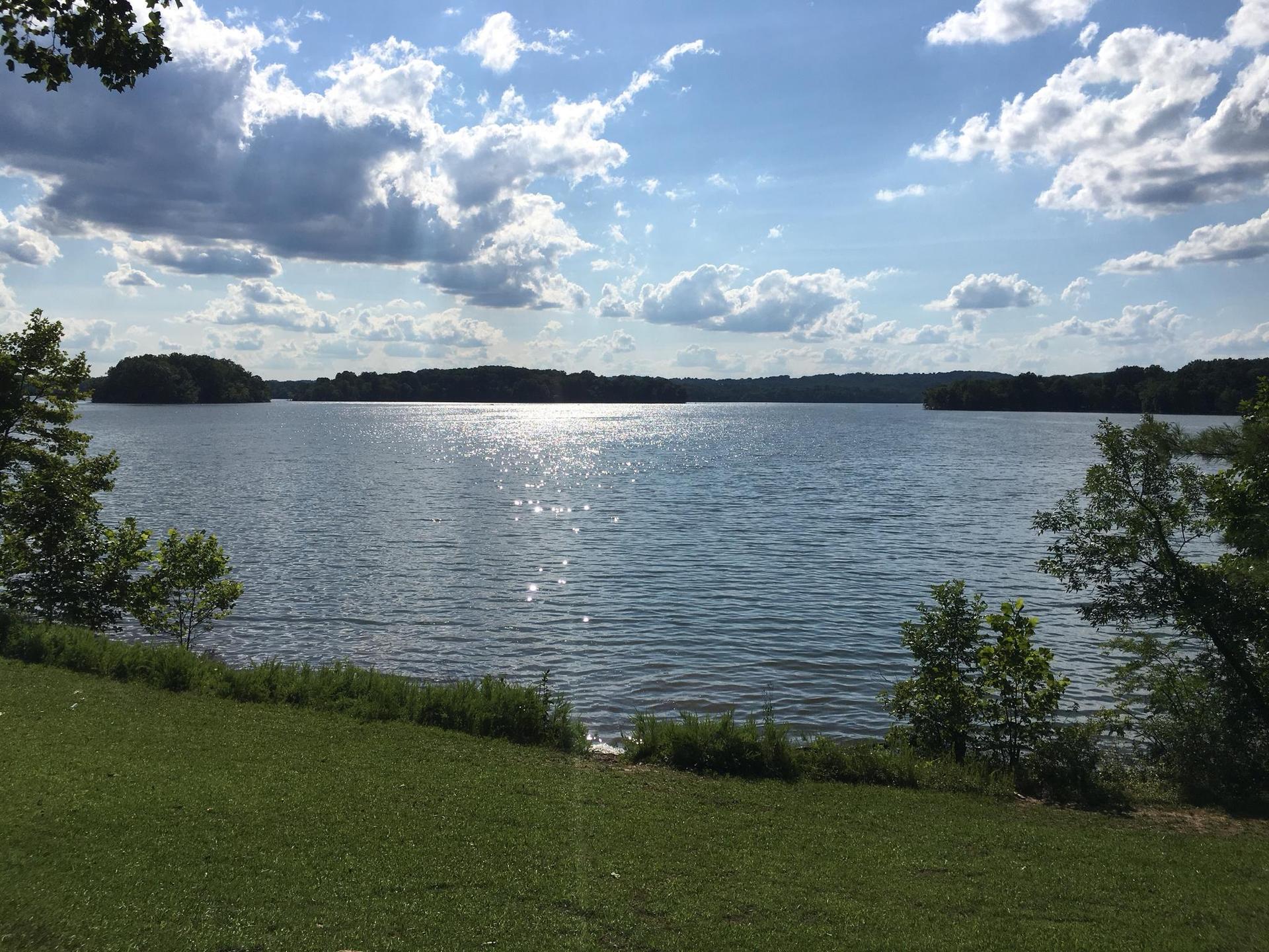 A wide lake with sun glinting on the water, surrounded by green shores.