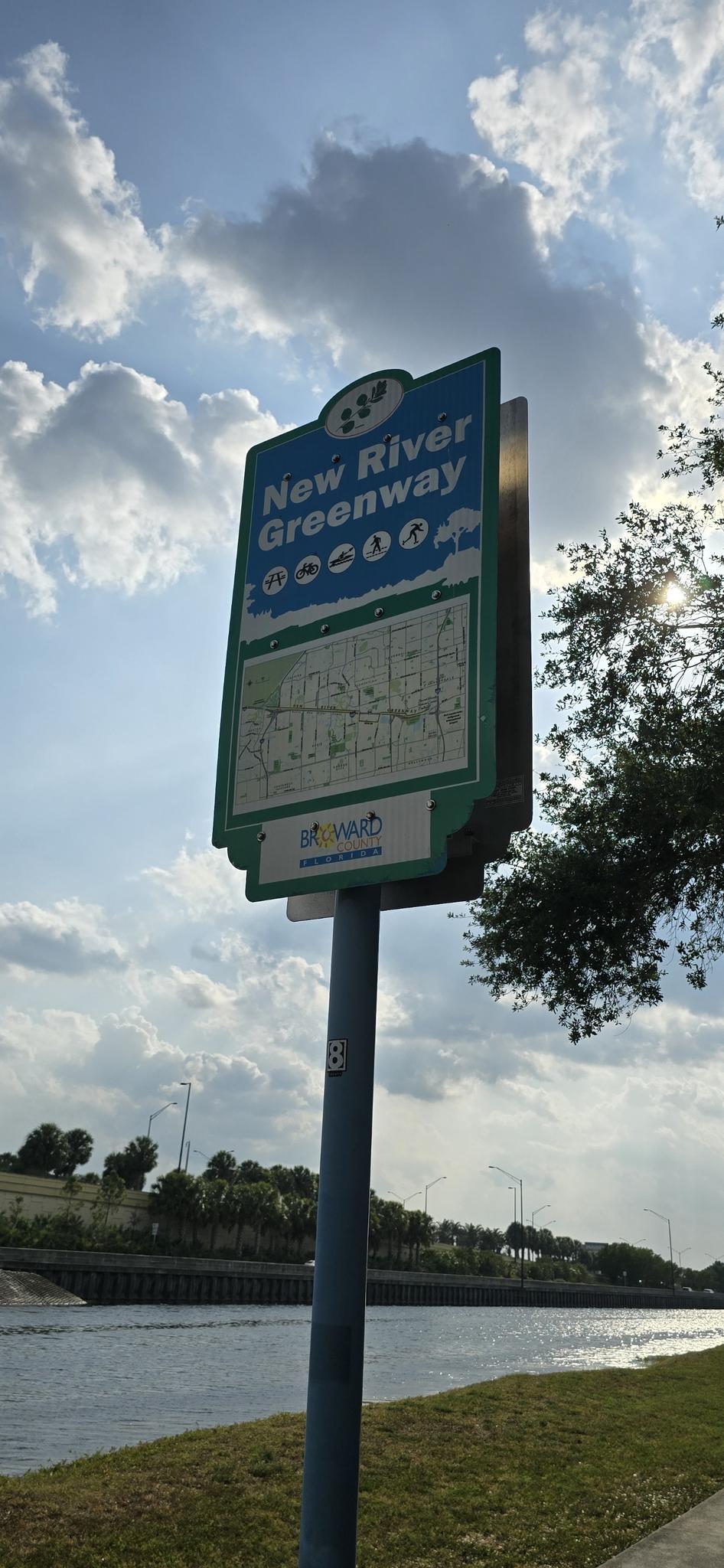 Tall blue and green sign next to a canal under a bright cloudy sky.