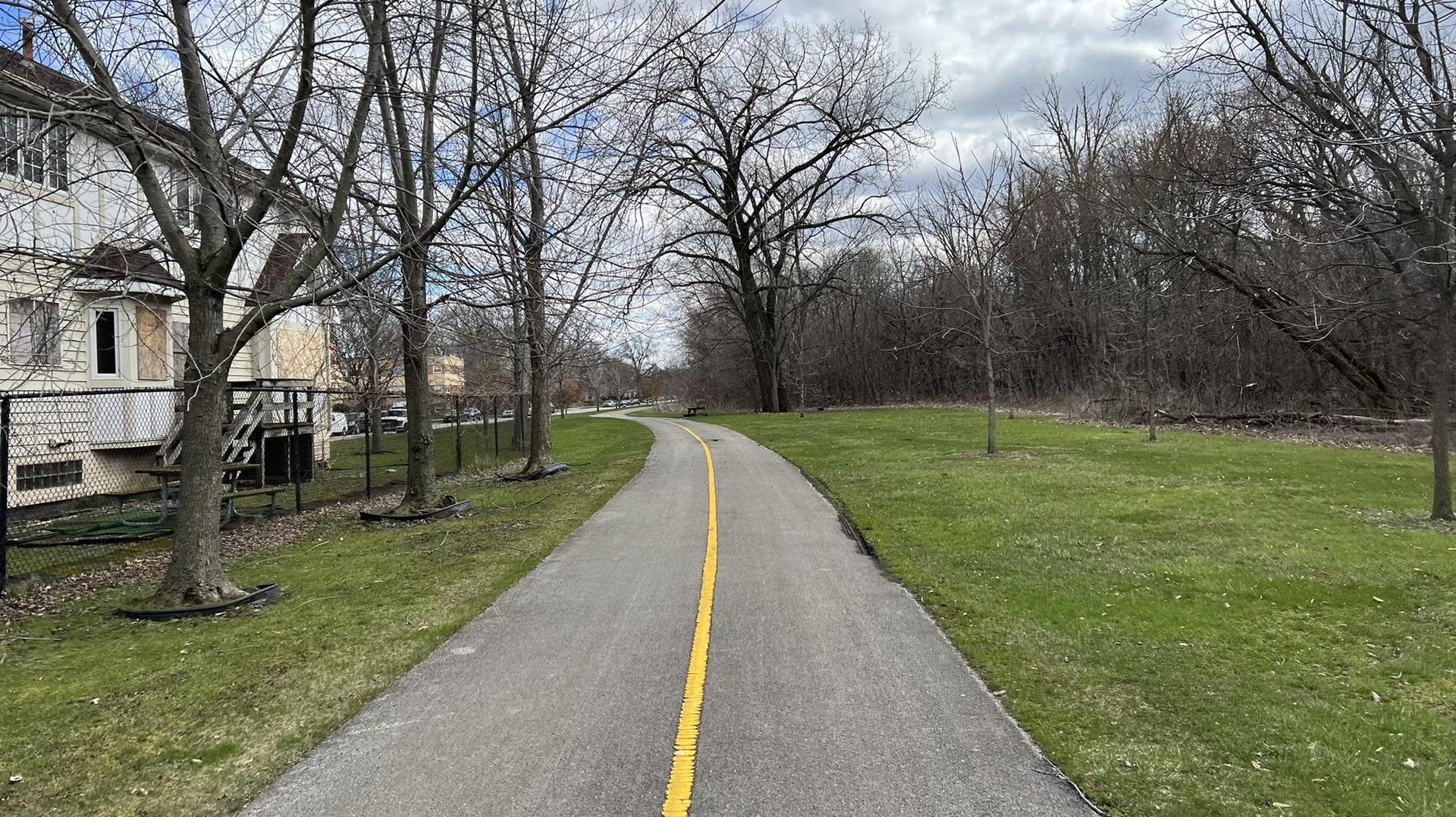Paved path with yellow line, bare trees, and buildings under cloudy sky.