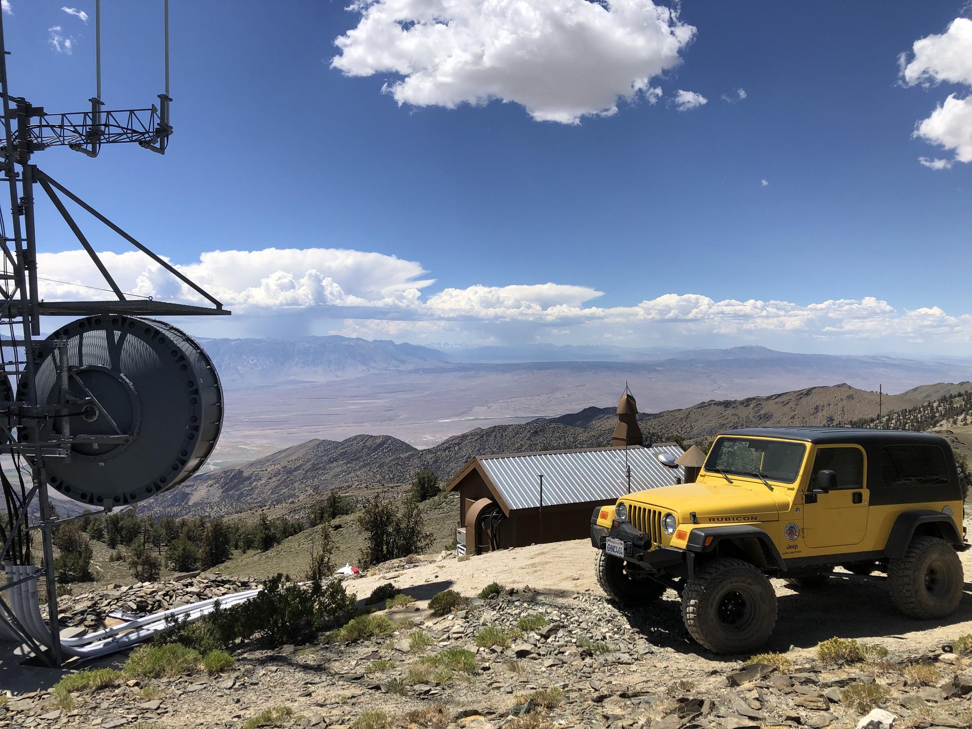 Yellow Jeep parked near a communication tower overlooking a vast mountain valley.