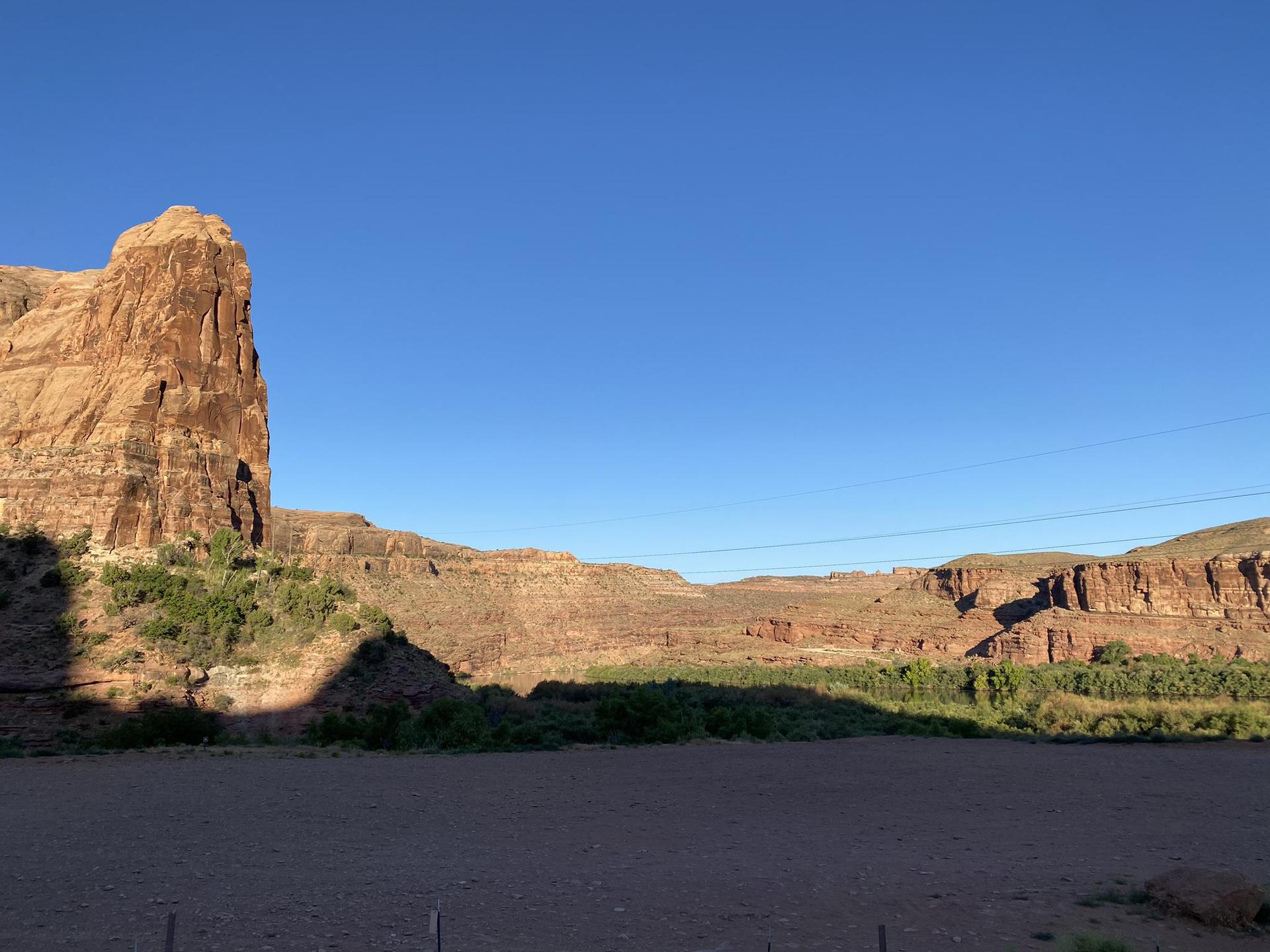 Tall red rock cliff casting a shadow over a green valley under a clear sky.