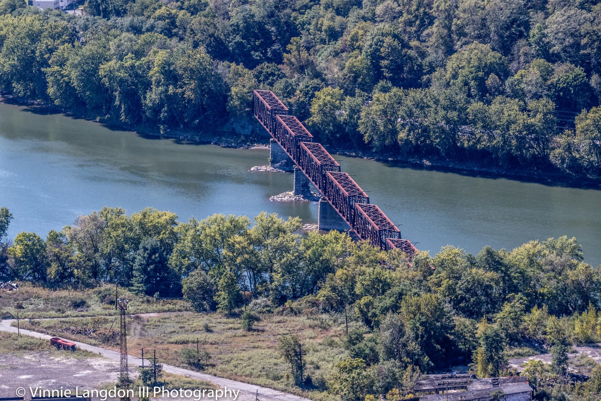 Aerial view of a long metal bridge crossing a wide river with green trees.