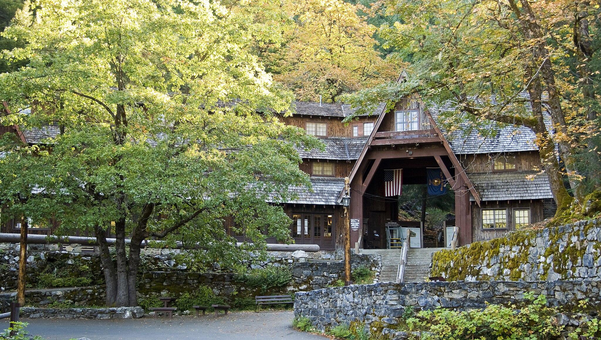 Rustic wooden lodge with an arched entrance surrounded by sunlit trees and stone walls.