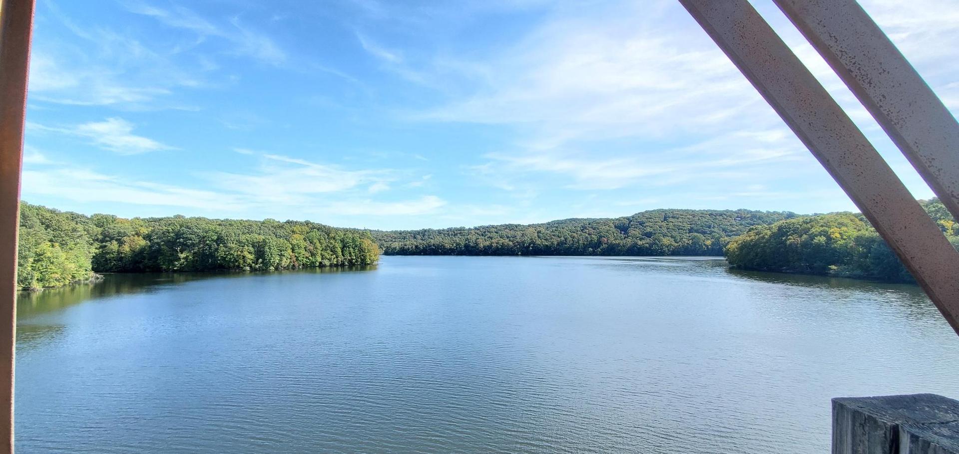 Expansive lake view from a bridge, surrounded by green hills, under a blue sky.