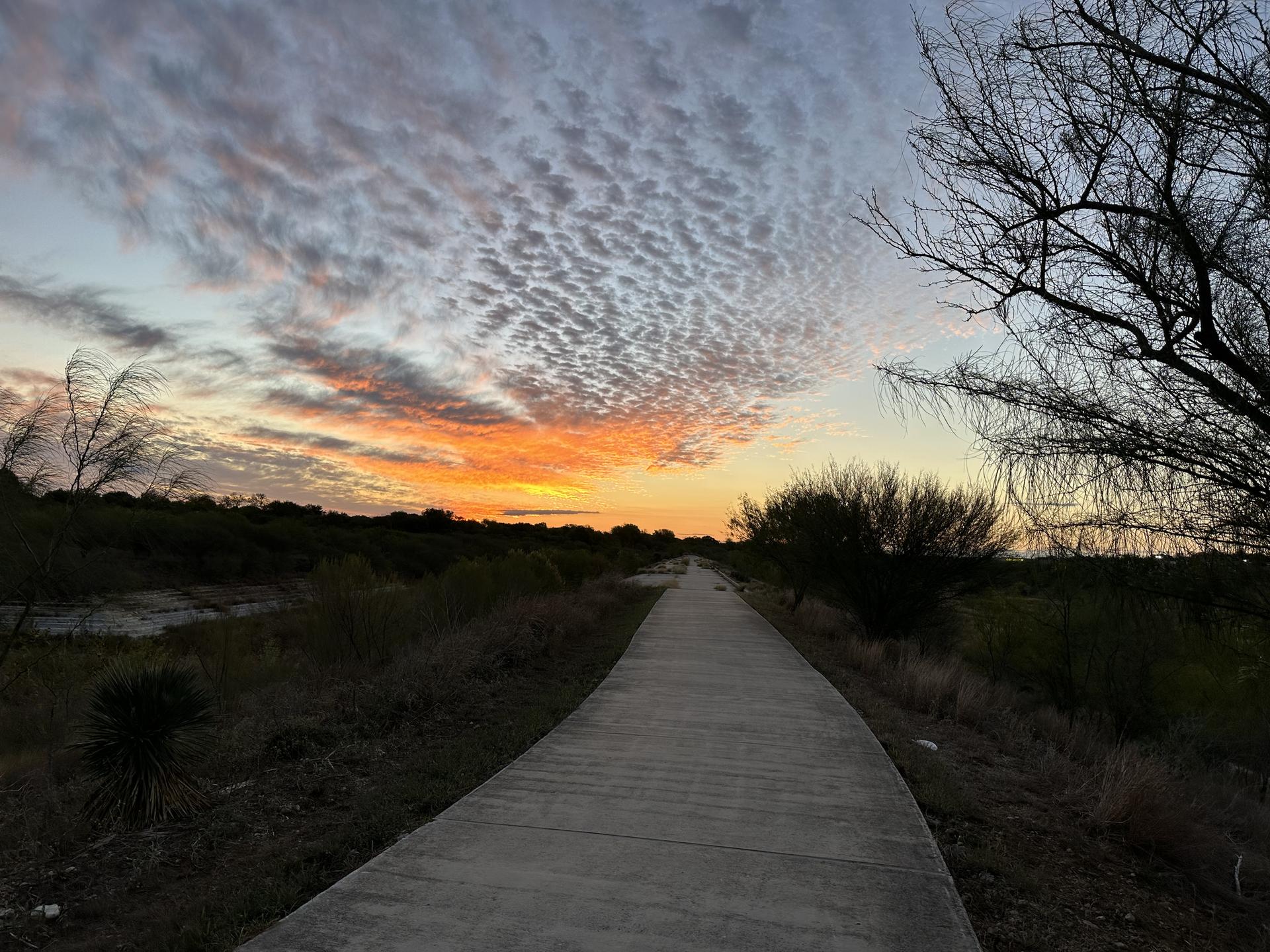 Paved path at sunset, vibrant orange and yellow clouds over a brushy landscape.