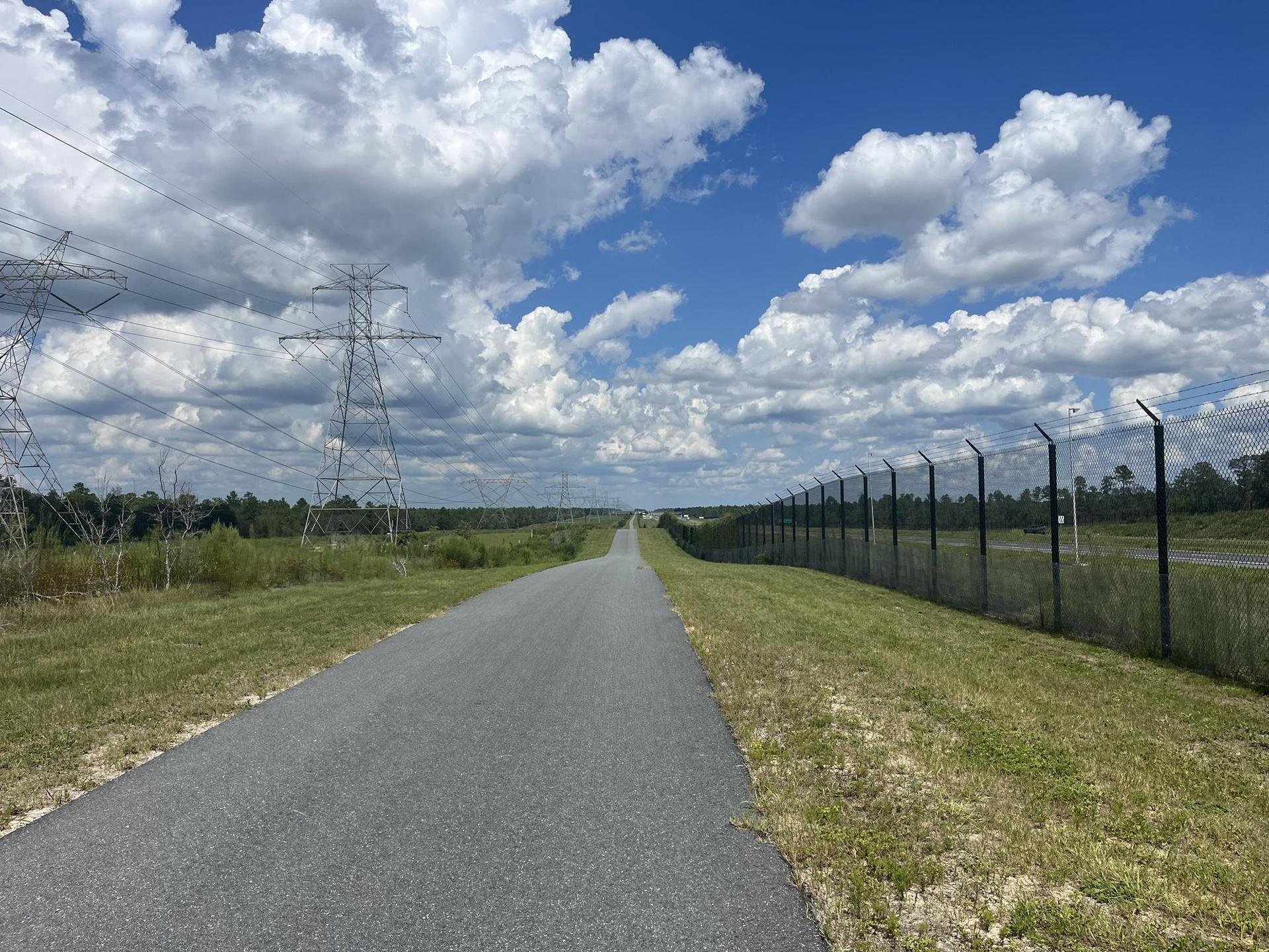 Paved road alongside a tall fence and power lines under a cloudy sky.