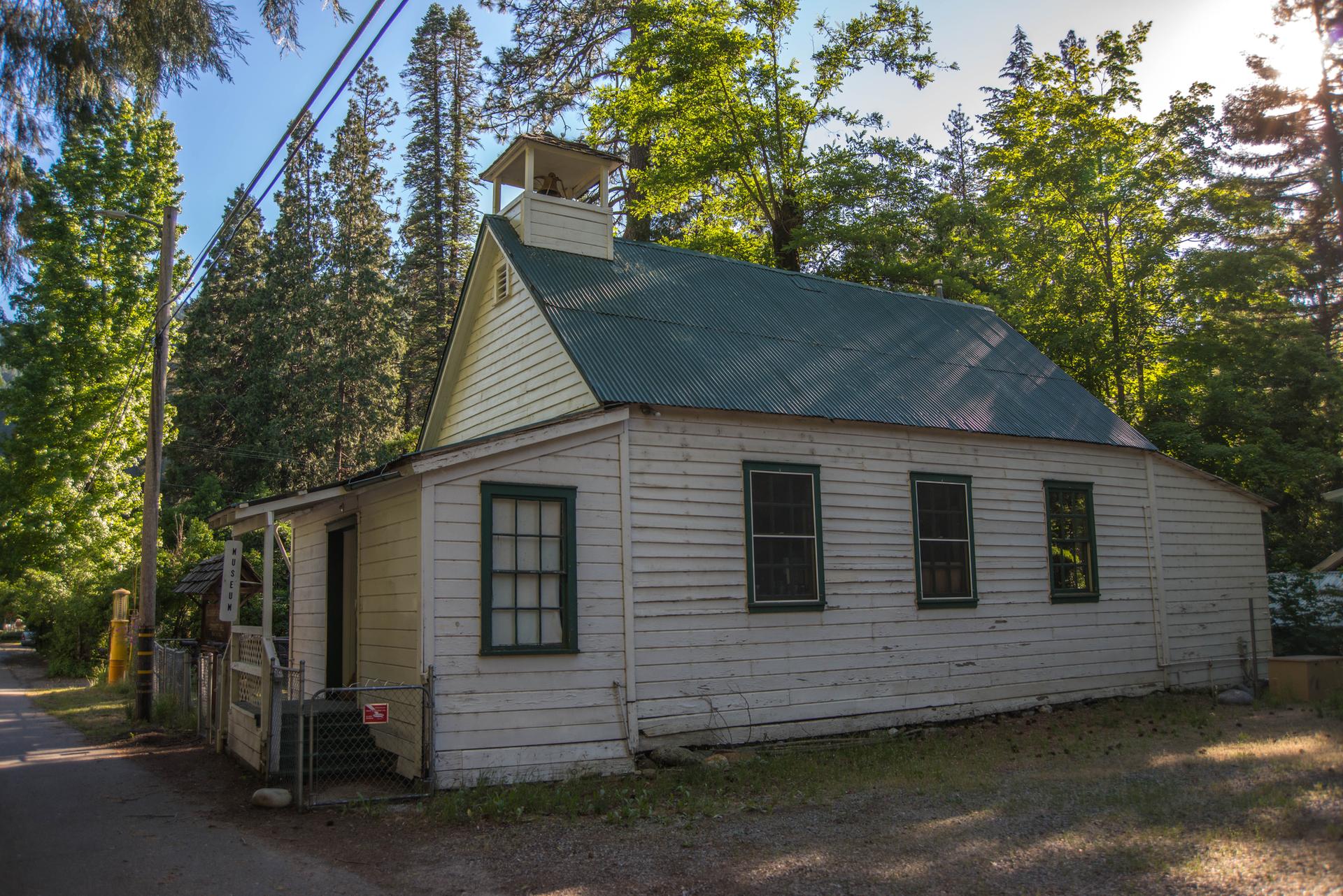 Old white wooden building with a green roof and bell tower, surrounded by trees.