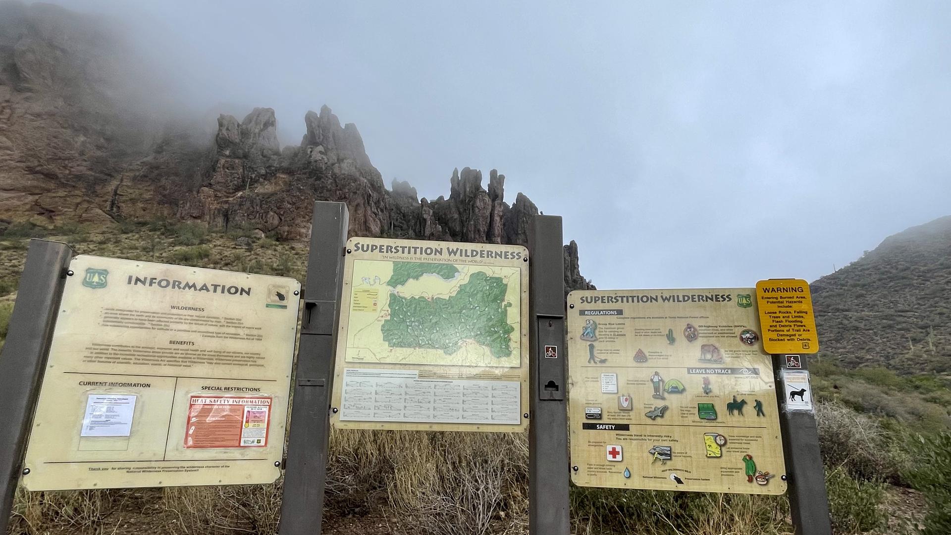 Three information signs stand before foggy, rugged mountains under an overcast sky.