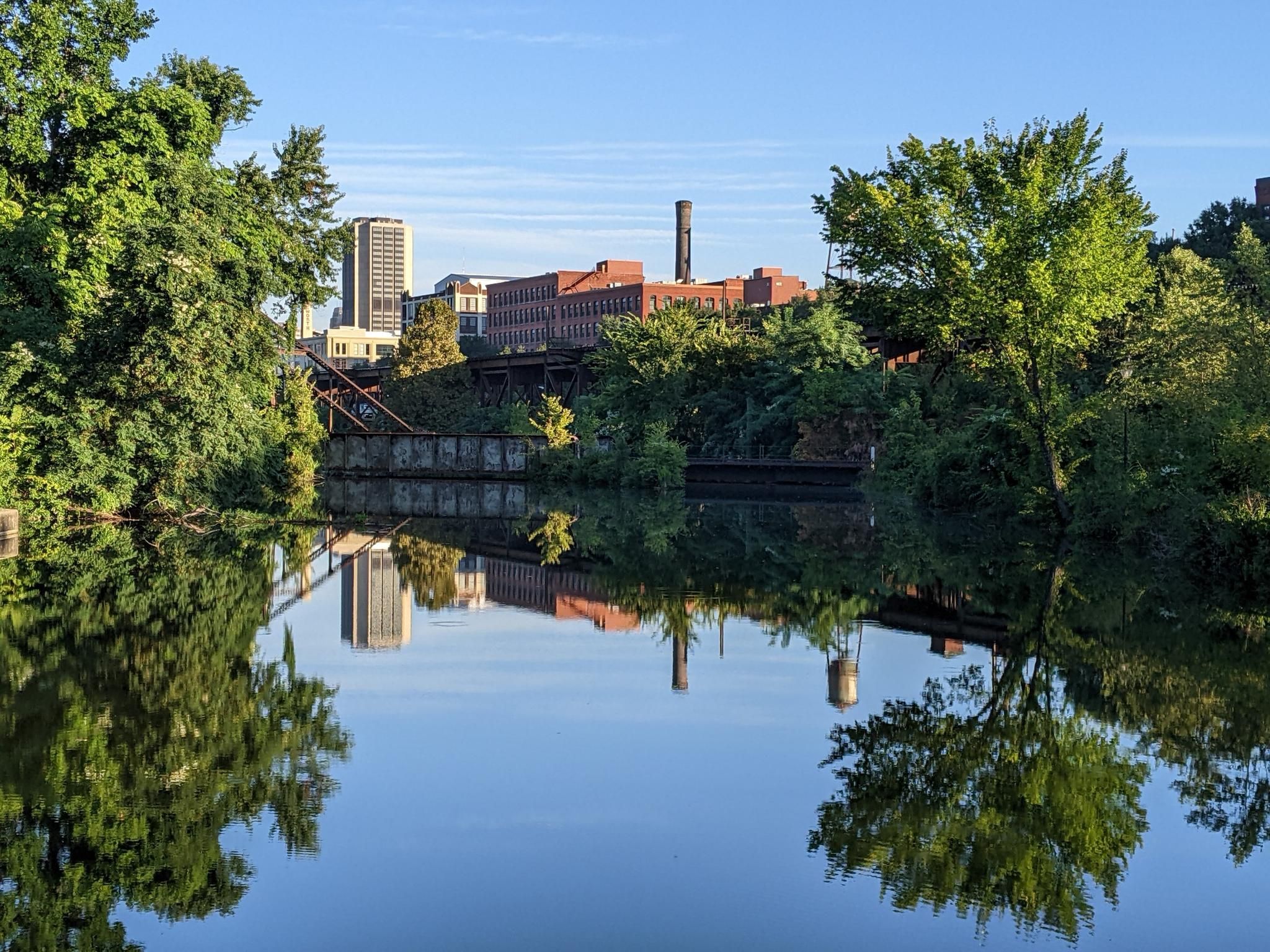 City buildings reflected in a calm river, surrounded by lush green trees. - UCI World Course (Zwift) – Dock Street Park loop from Richmond
