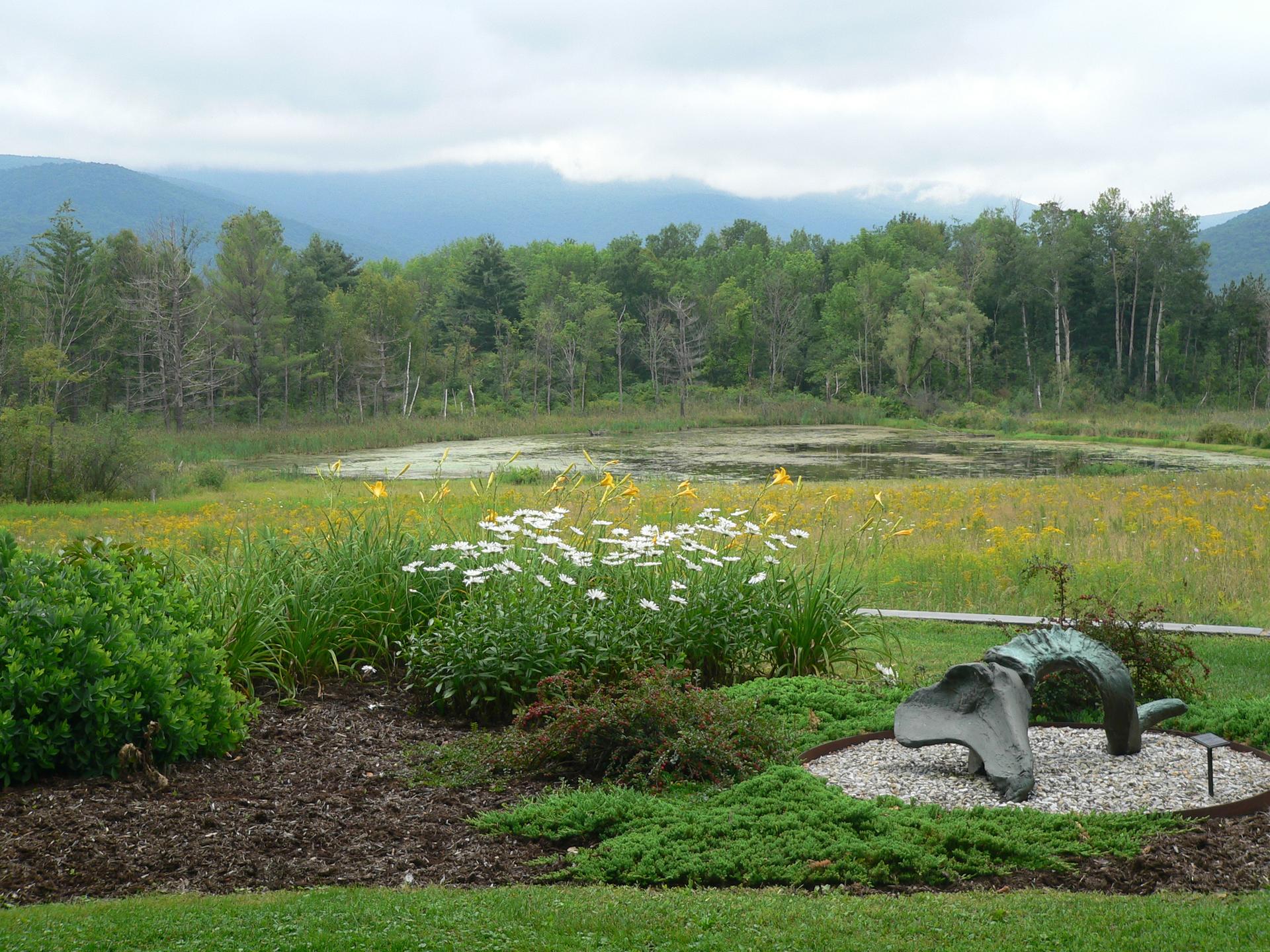 Pond, wildflowers, forest, and mountains under cloudy sky, with a sculpture.