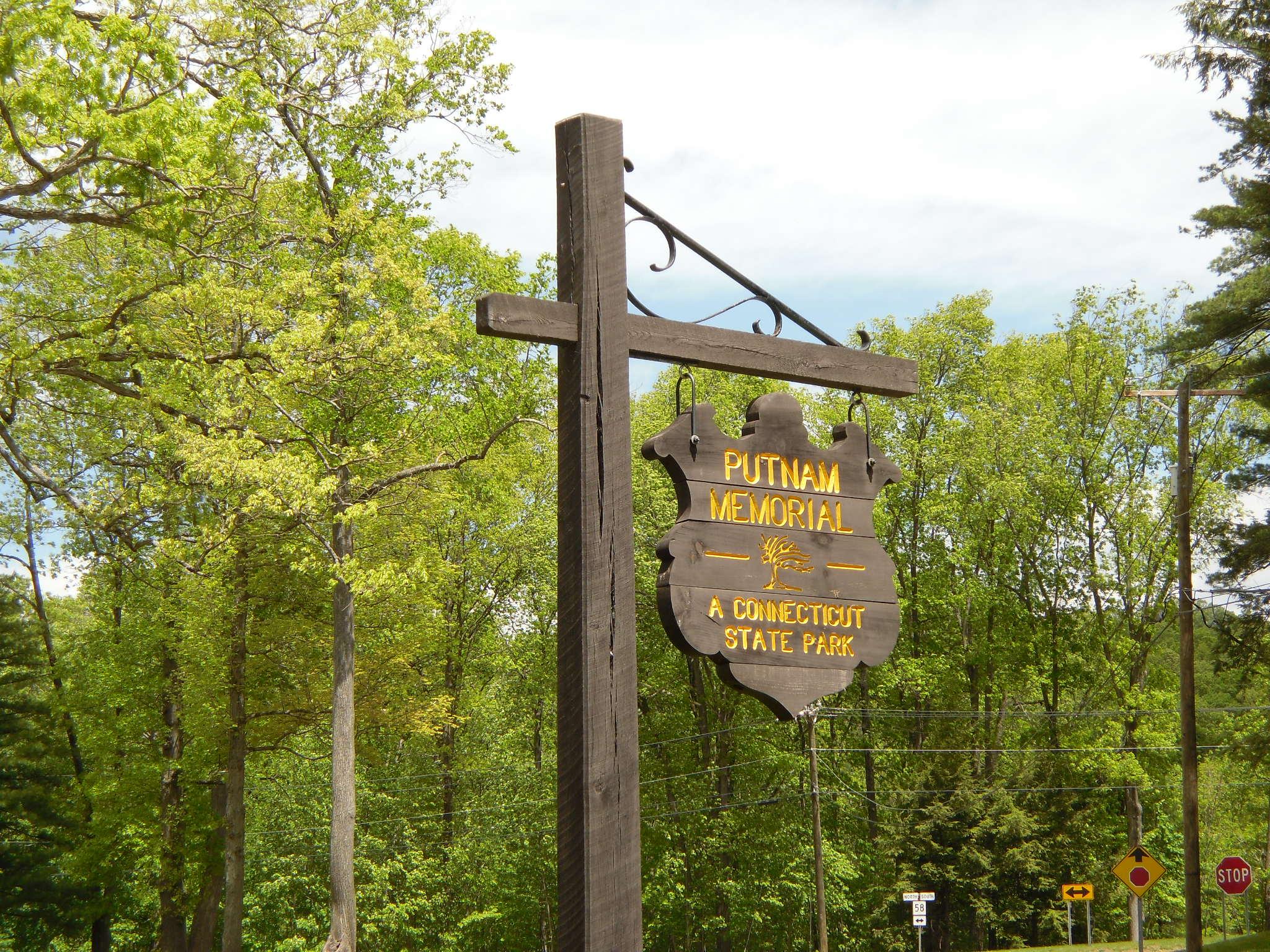 Wooden sign on a post, surrounded by lush green trees in bright daylight.