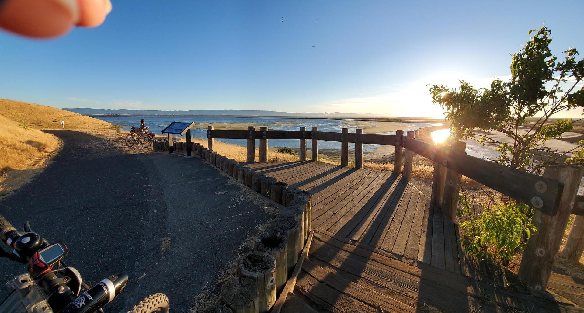 Golden hour over a wooden boardwalk by water, with a person on a bike.