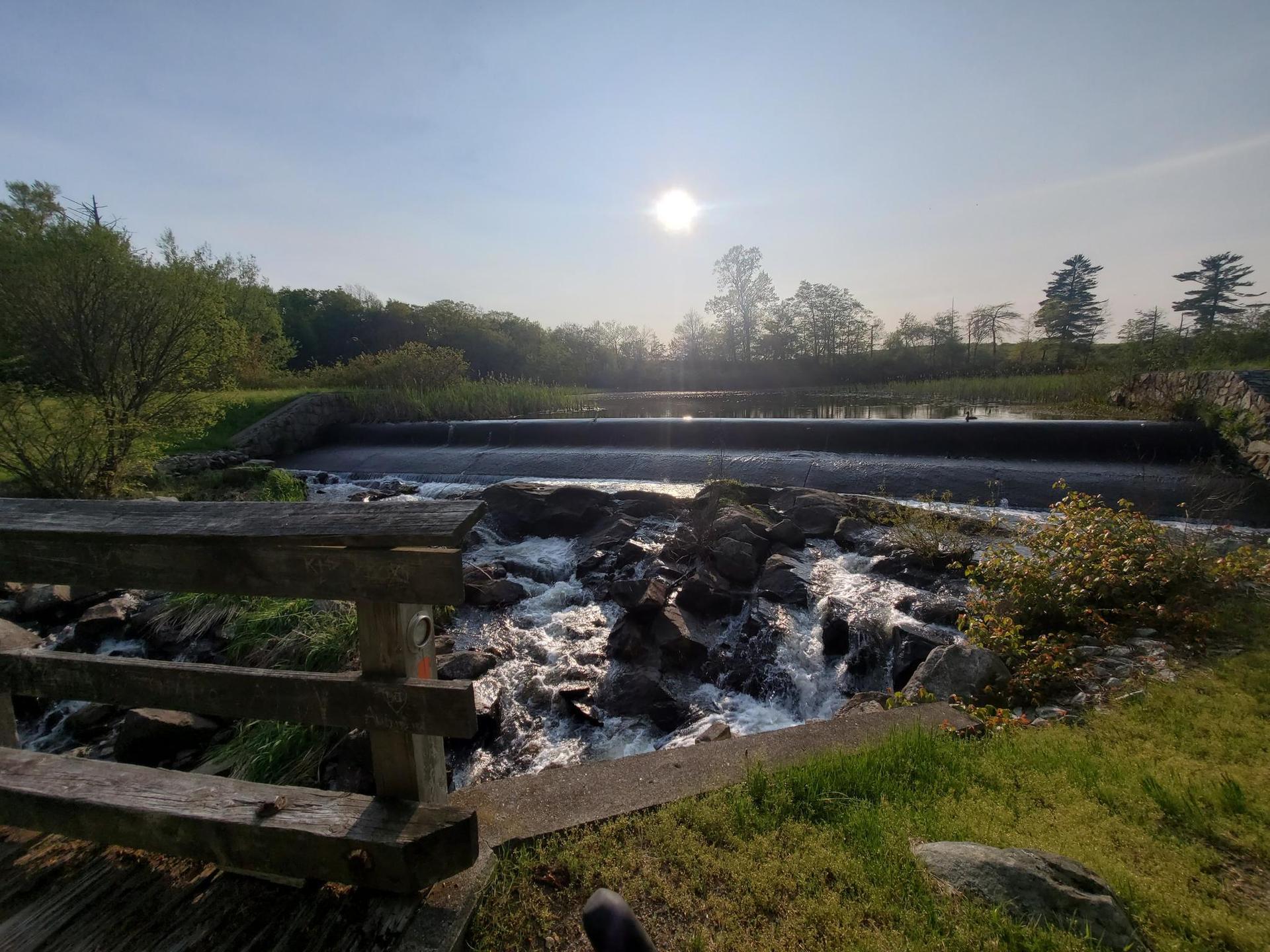 Sunlight glints over a waterfall and dam, with a wooden railing foreground.