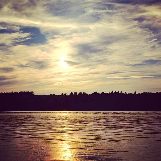 Bright sun reflects on rippling water with a dark forested horizon under cloudy sky.