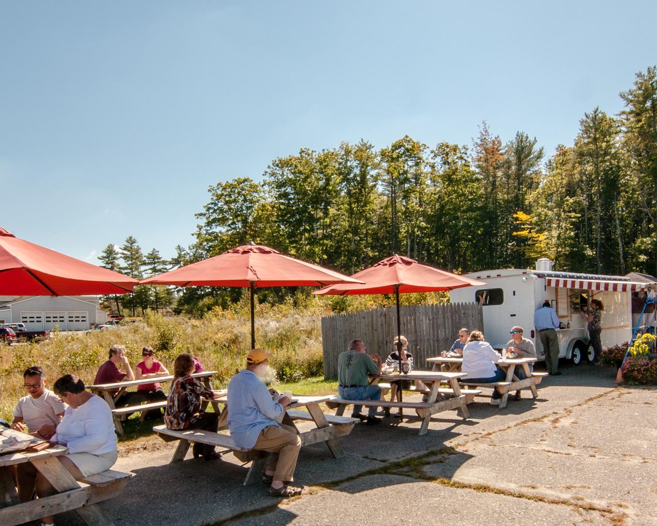 People eating at picnic tables with red umbrellas on a sunny day.