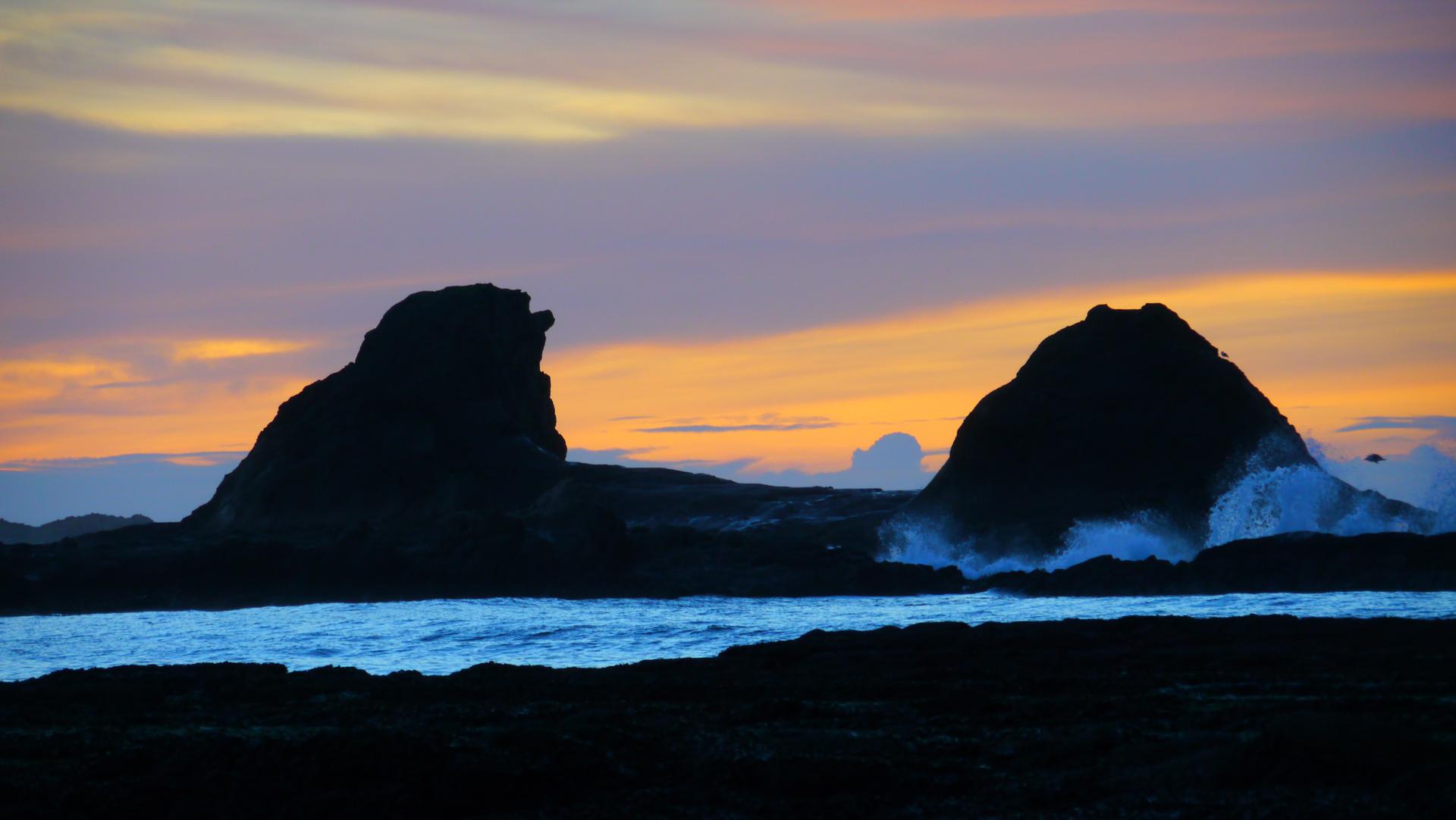 Two silhouetted rock formations against a vibrant sunset sky with crashing waves.