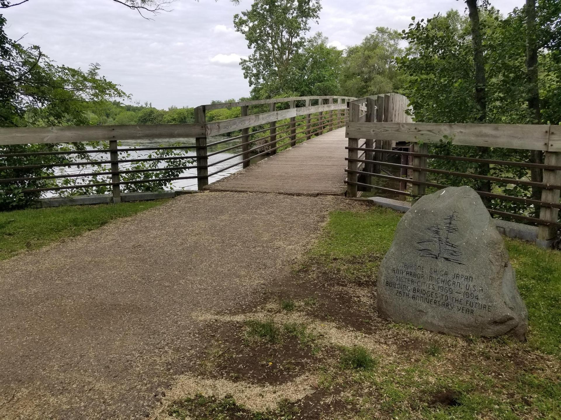 A gravel path leads to a wooden bridge over water, with a large rock.