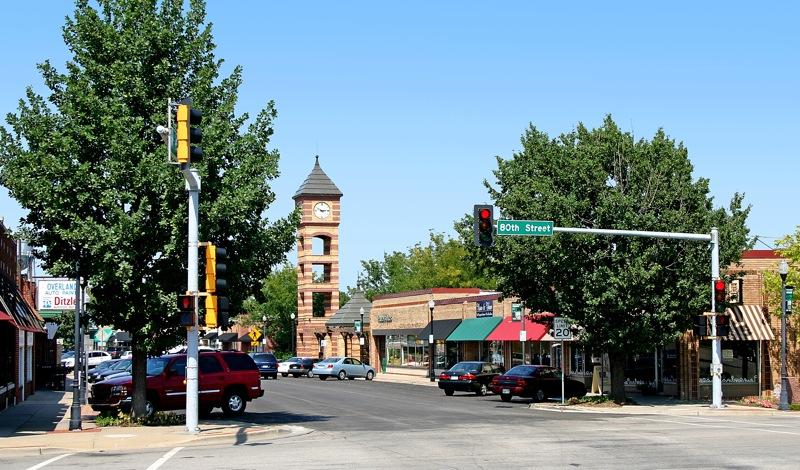 A sunny town intersection with cars, buildings, and a prominent clock tower.