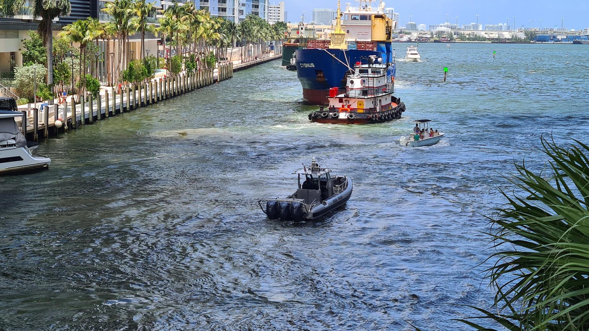 Boats navigate a waterway, with a large ship and tugboat in the background.