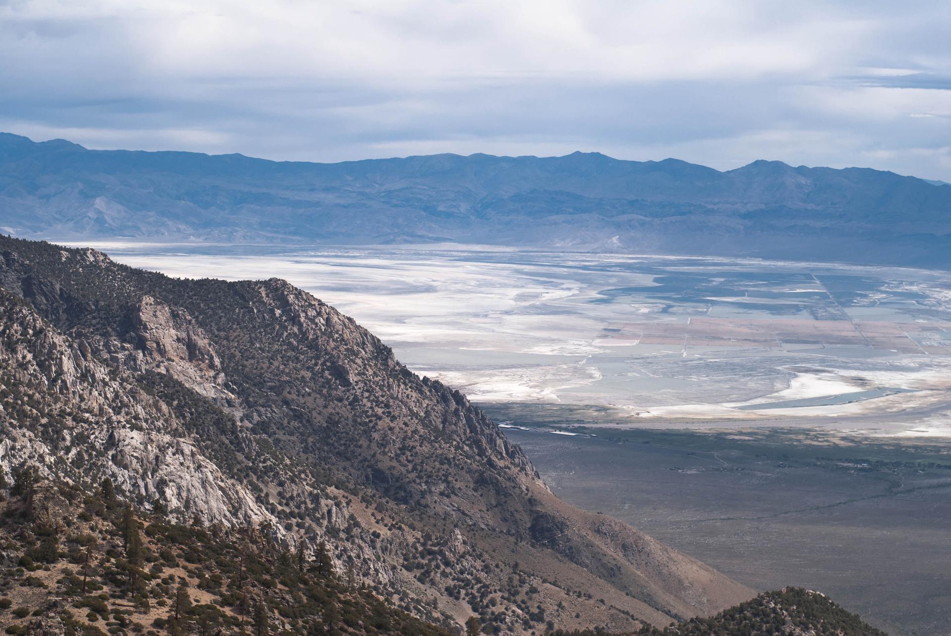 Vast, dry valley with salt flats seen from a rocky mountain slope under cloudy skies.