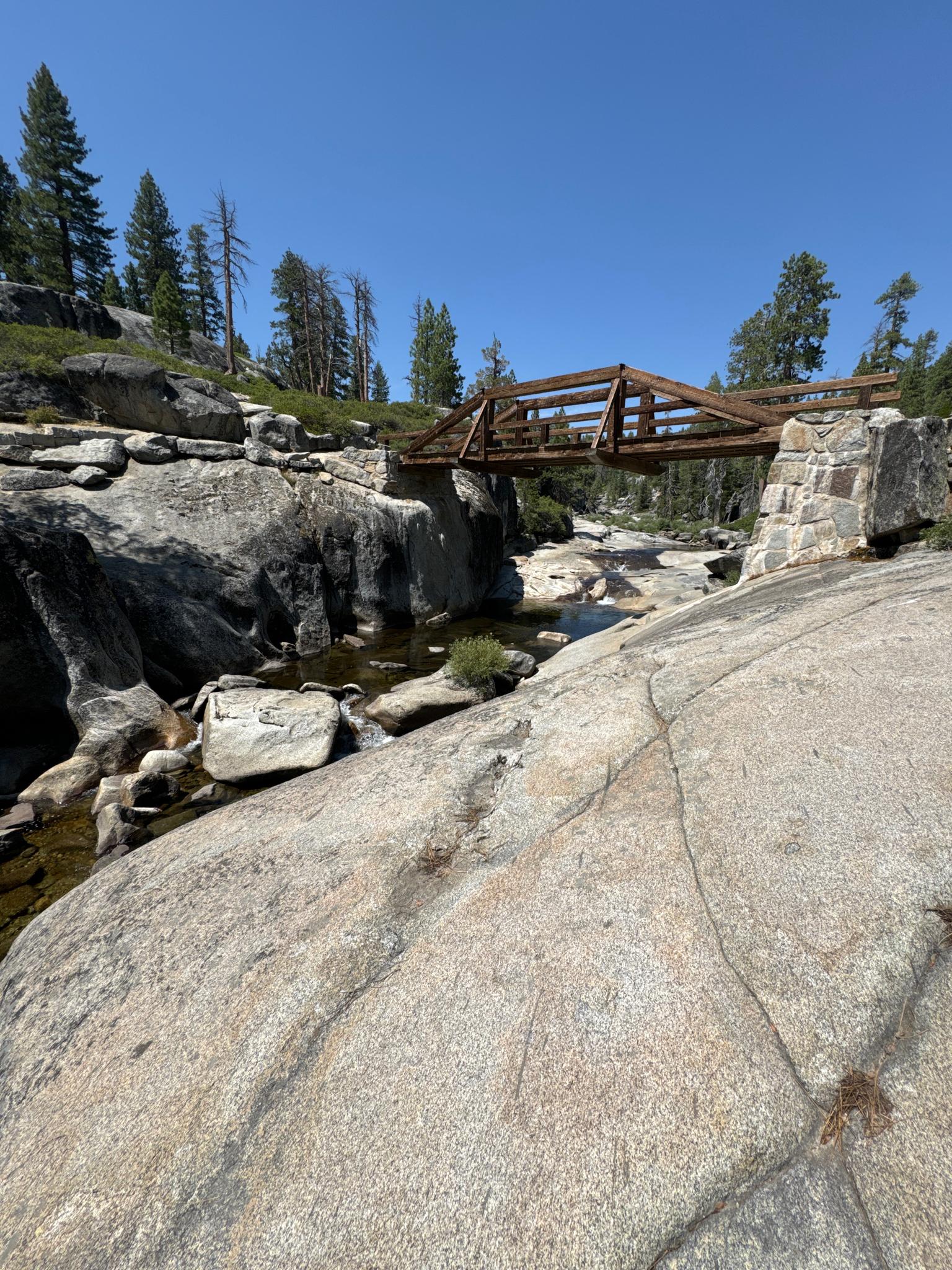 Rustic wooden bridge spans a rocky stream under a clear blue sky.