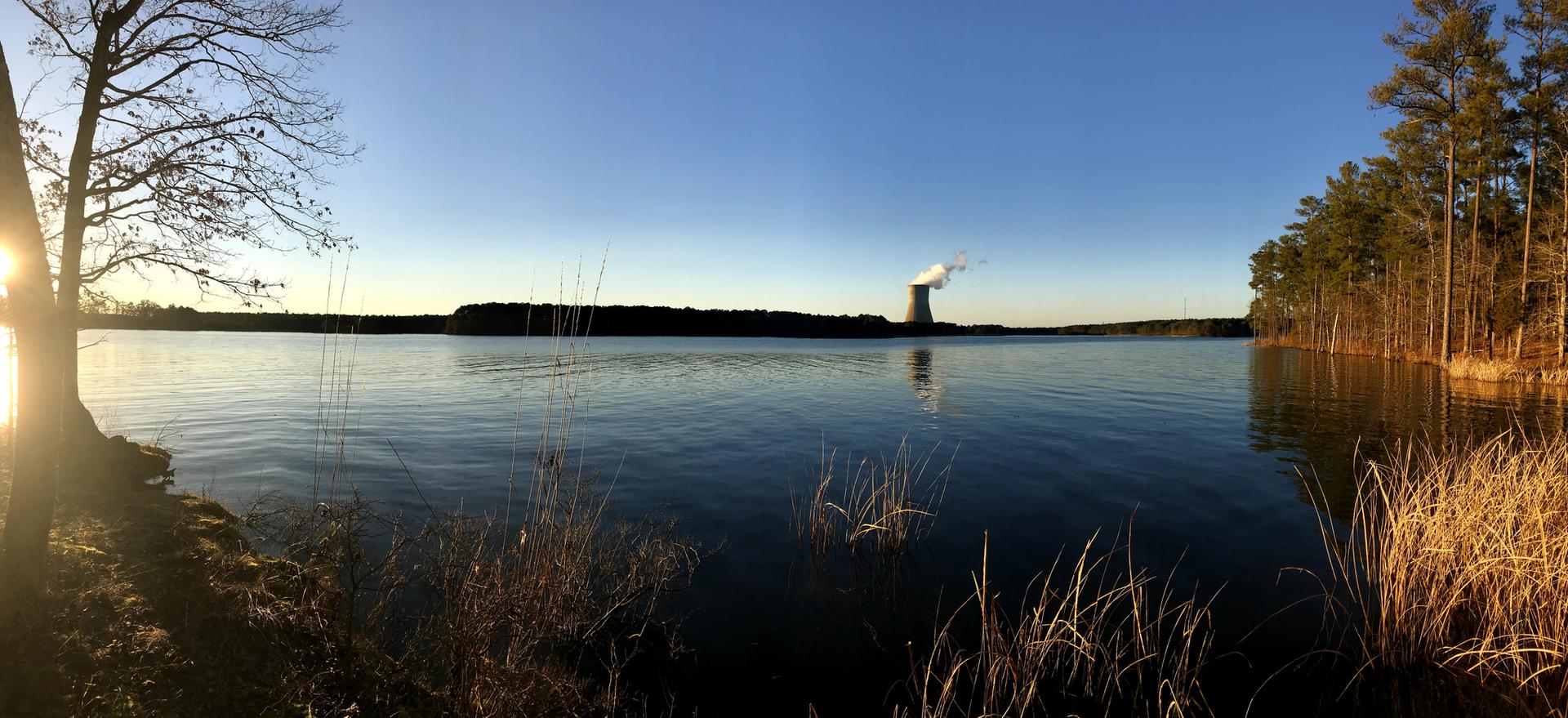 Wide lake with distant cooling tower under clear sky, sun on left.