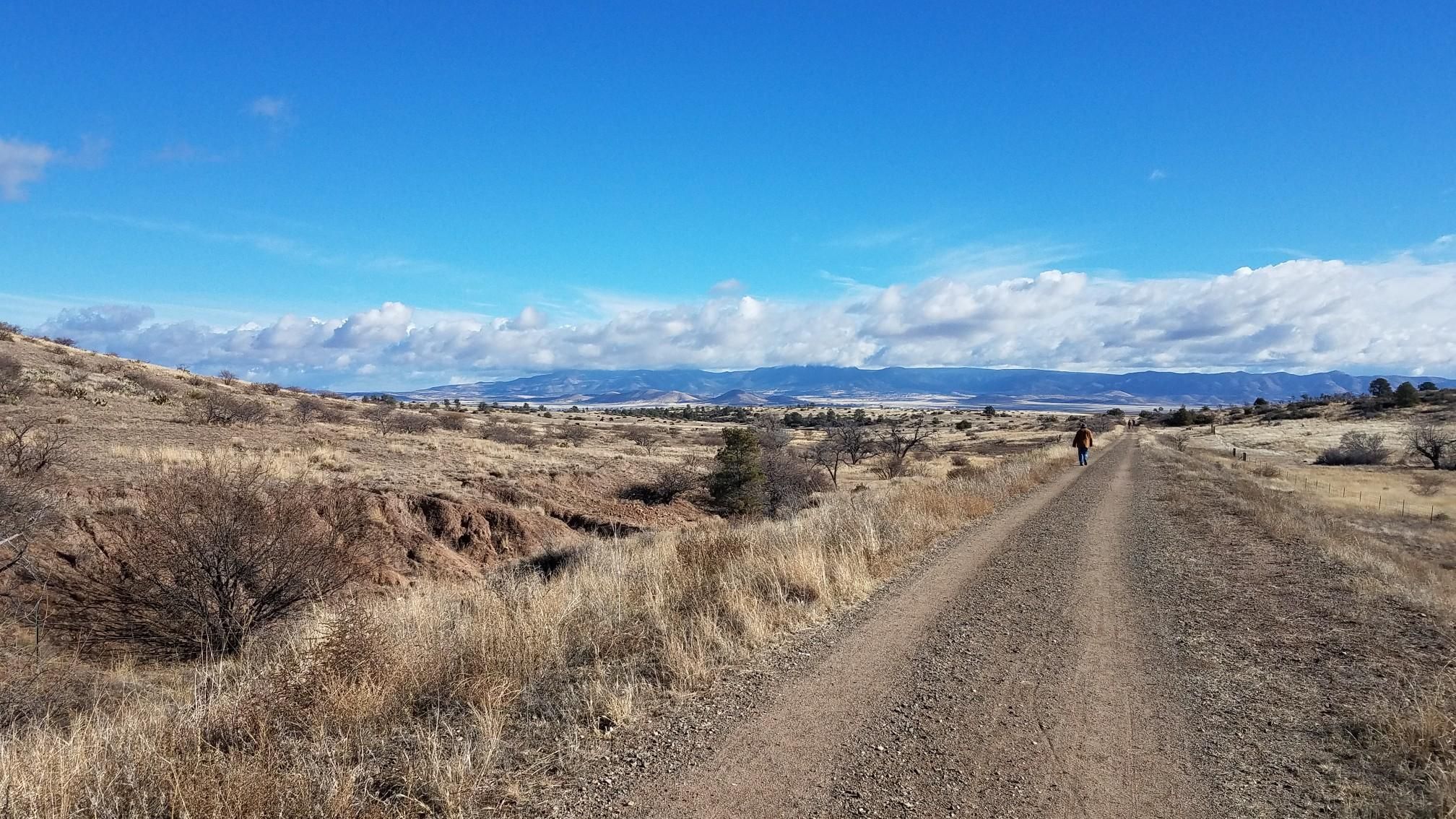 A person walks on a dirt path through a dry, expansive landscape under a blue sky. - Peavine Trail – Iron King Trail loop from Granite Dells