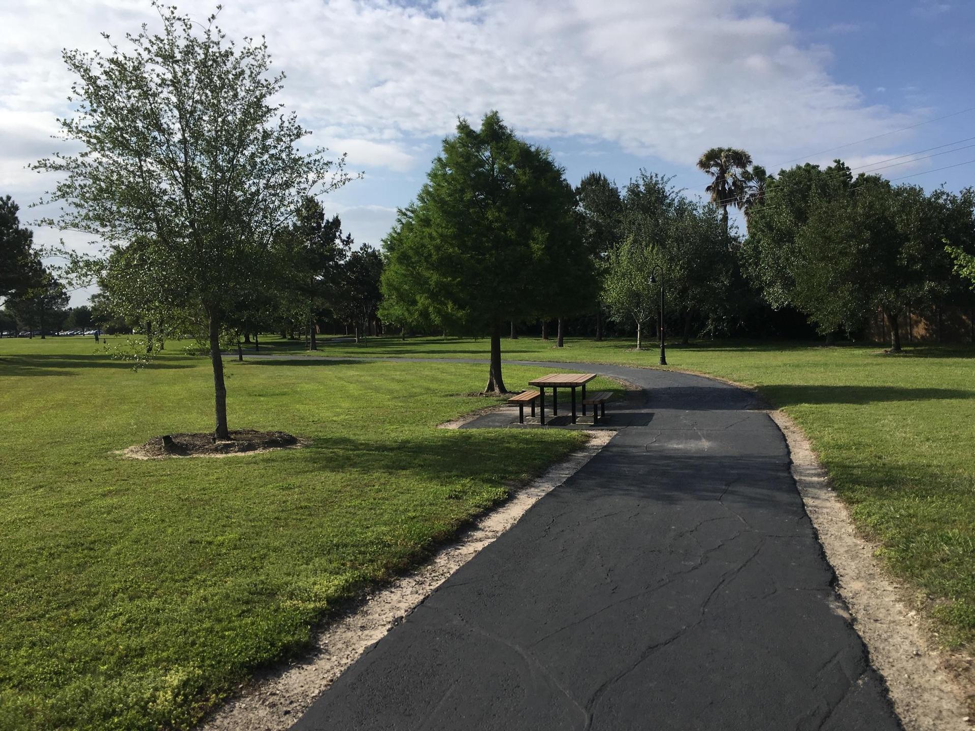 A paved path winds through a grassy park with trees and a picnic table.