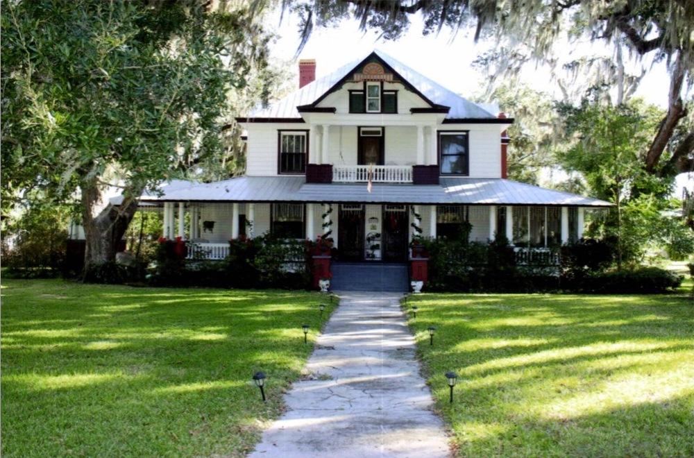 Grand white house with wide porch and green lawn under mossy trees.
