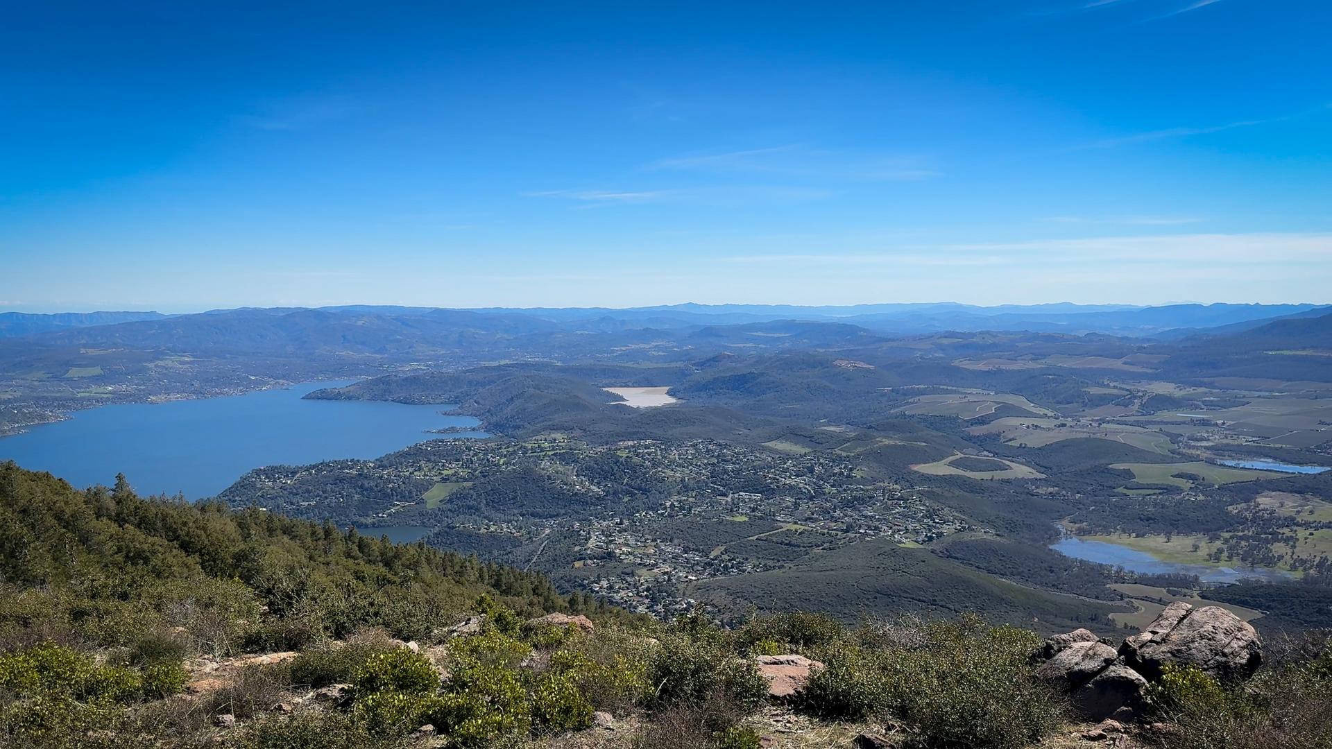 Panoramic view of a large lake, town, and mountains under a clear blue sky.
