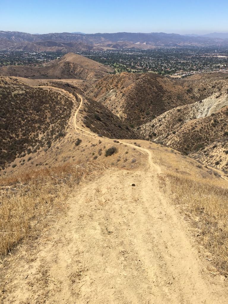 A winding dirt trail descends through dry, arid hills under a clear sky.
