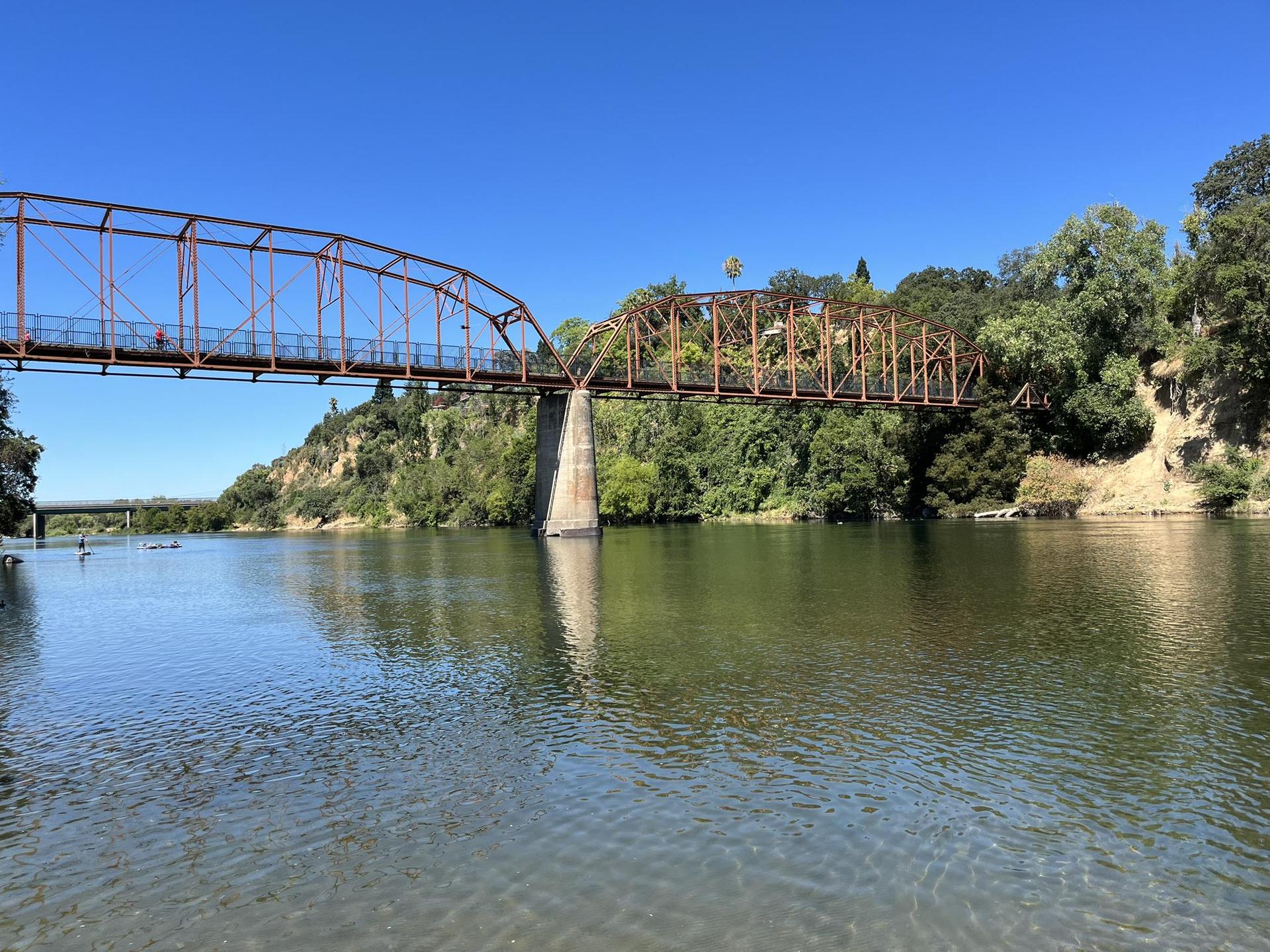 Orange truss bridge spans a wide river with green, tree-lined banks.