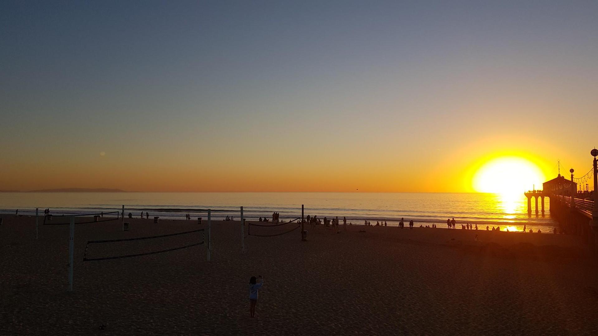 Golden sunset over a busy beach with a pier and many people.
