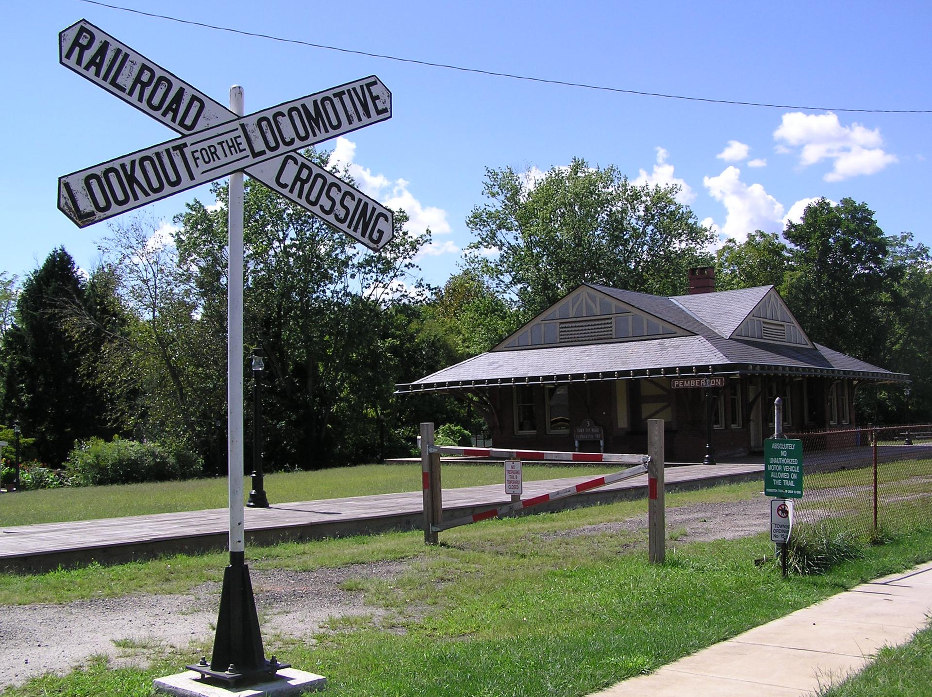 Railroad crossing sign in foreground, old station building under bright sky.