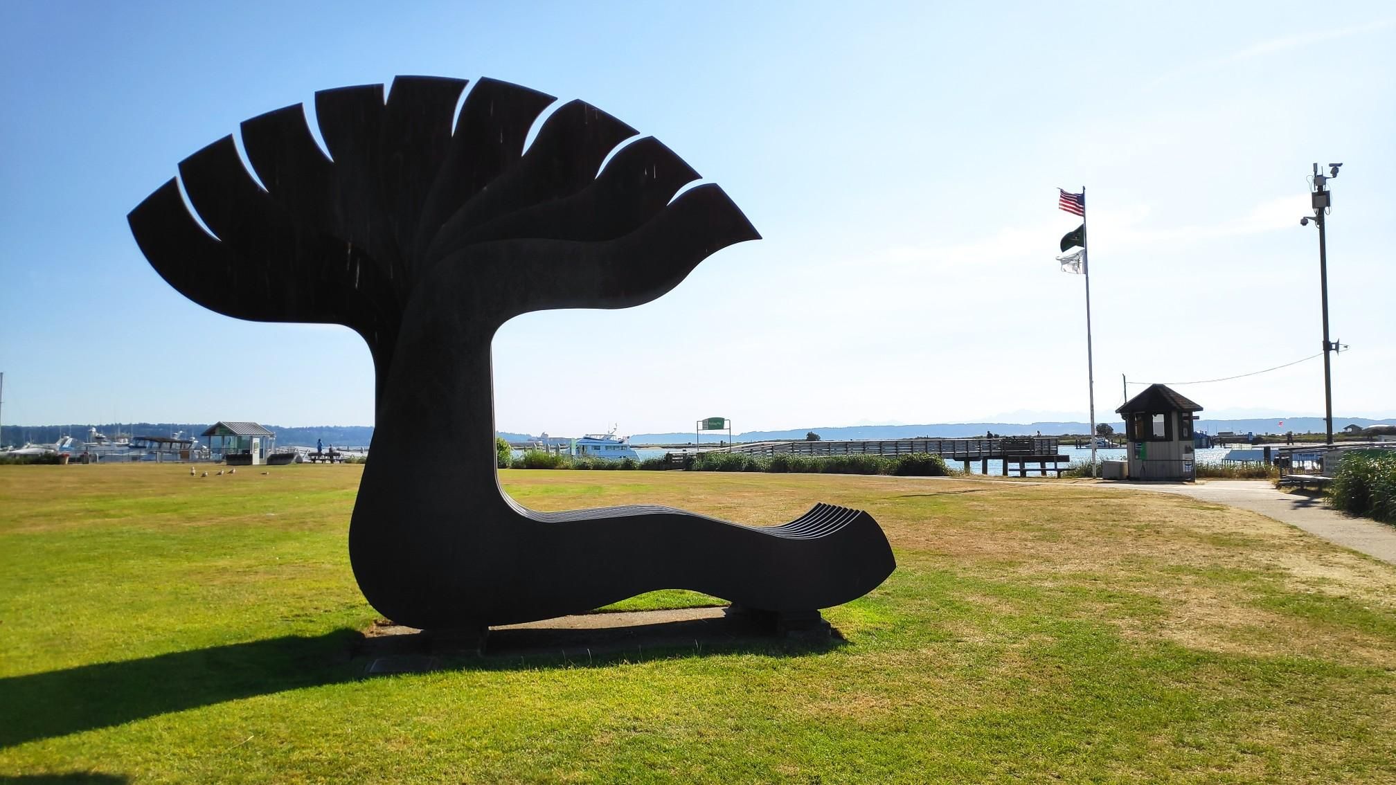 Large dark abstract sculpture on grass, with water and flags in background. - Hiking loop from Everett