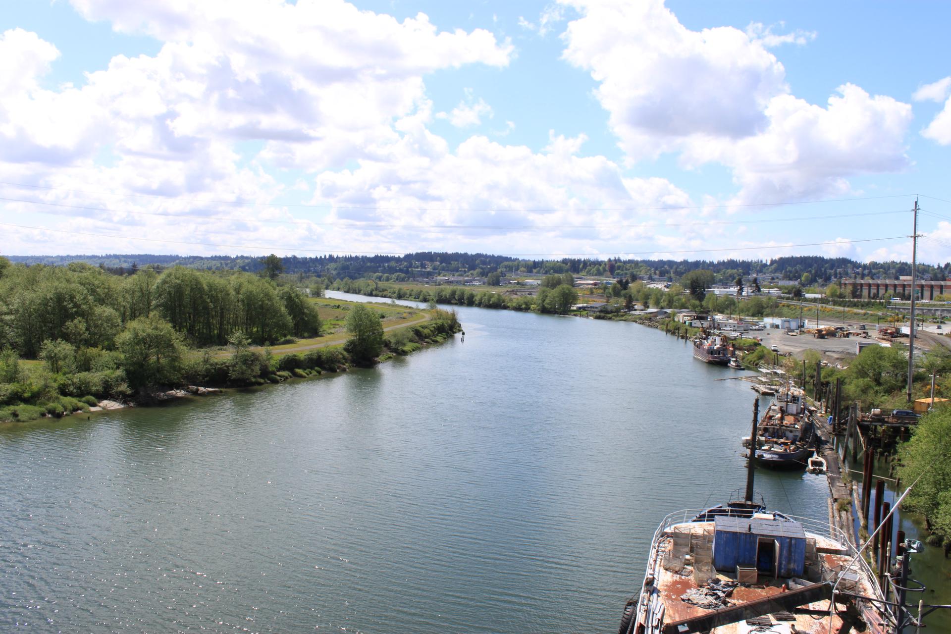 Wide river with green banks, old boats docked, under a bright, cloudy sky.