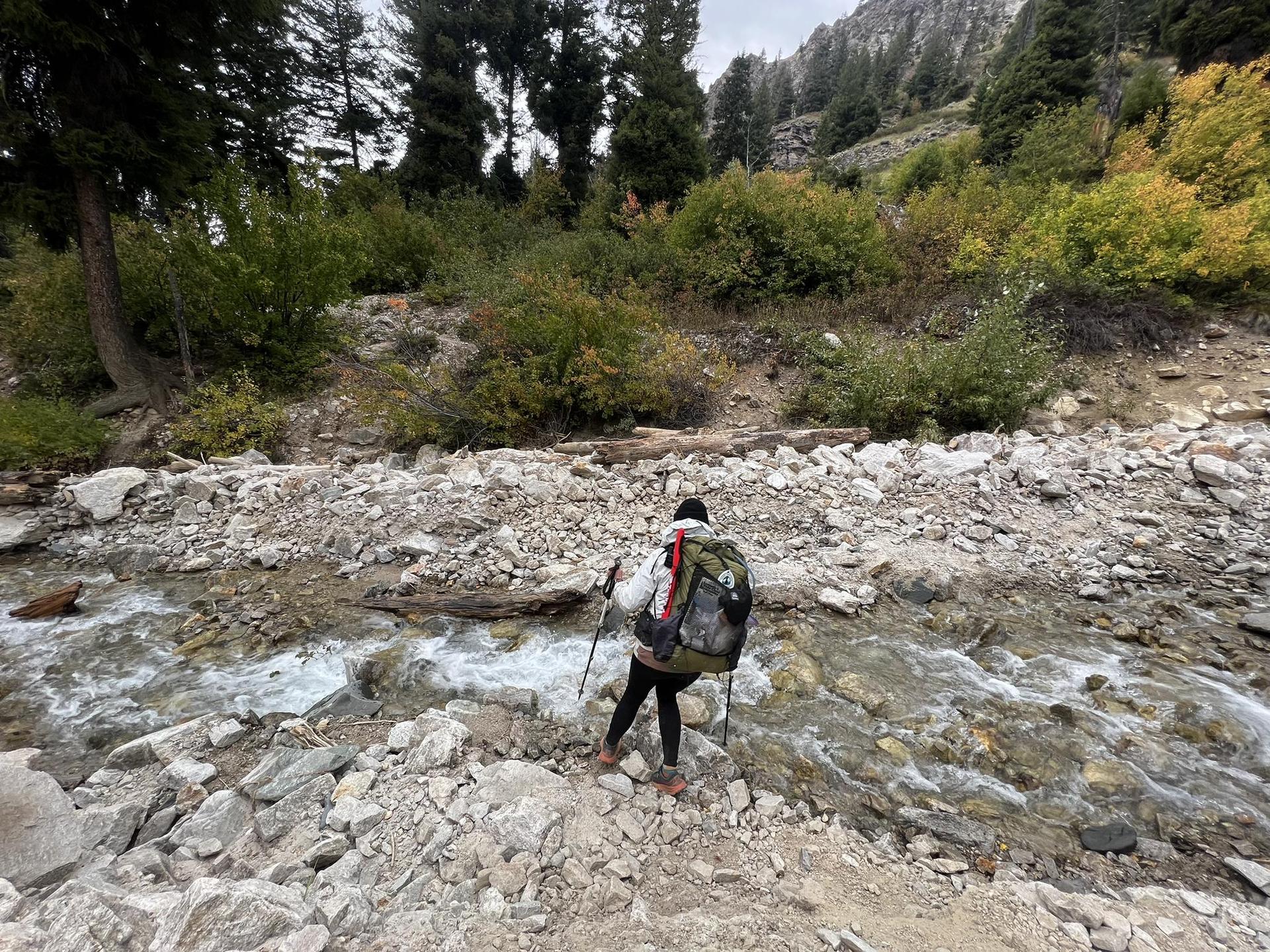 Hiker with backpack and poles crossing a rocky stream in a forested landscape.