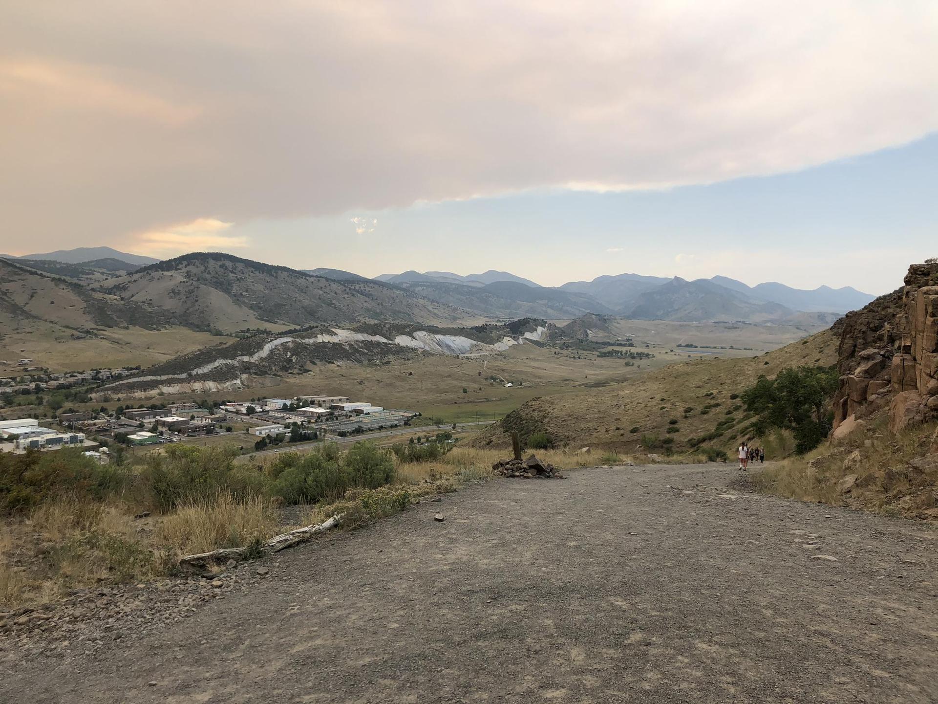 Gravel path overlooking a valley with distant town and mountains under cloudy skies.