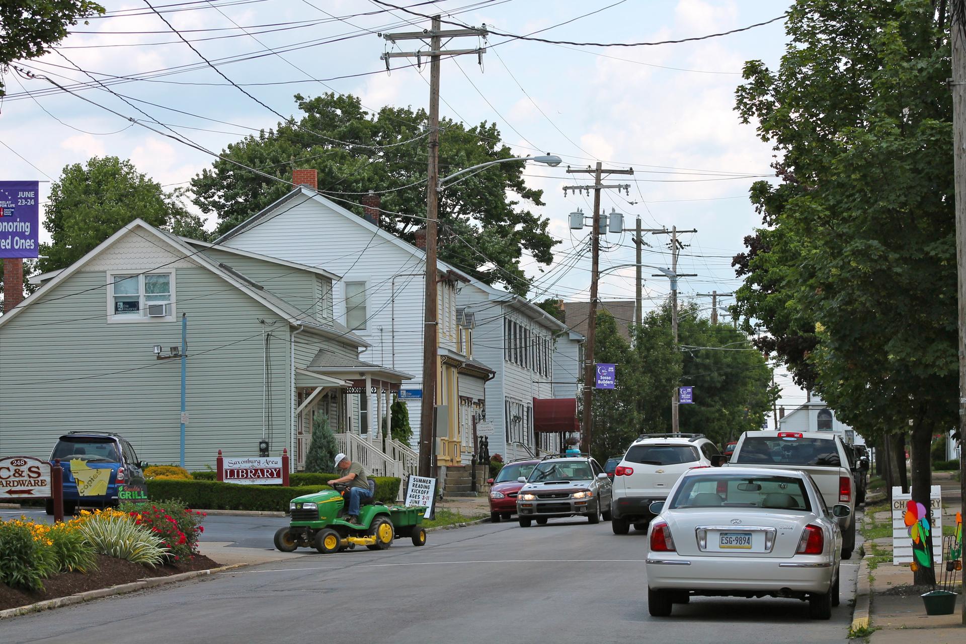 A person drives a lawnmower on a street with houses and cars.