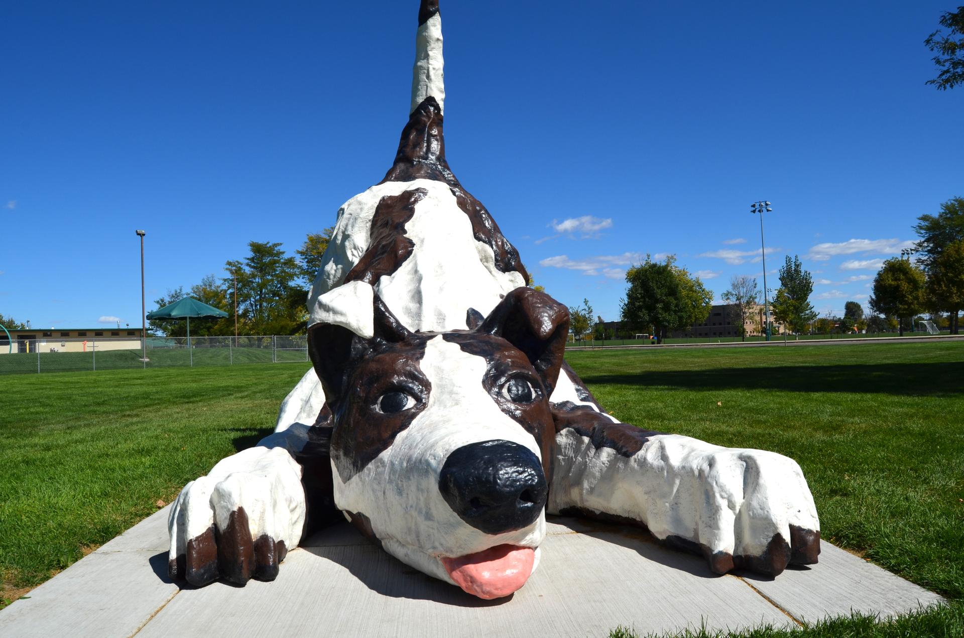 Large black and white dog statue on concrete in a sunny grassy field.