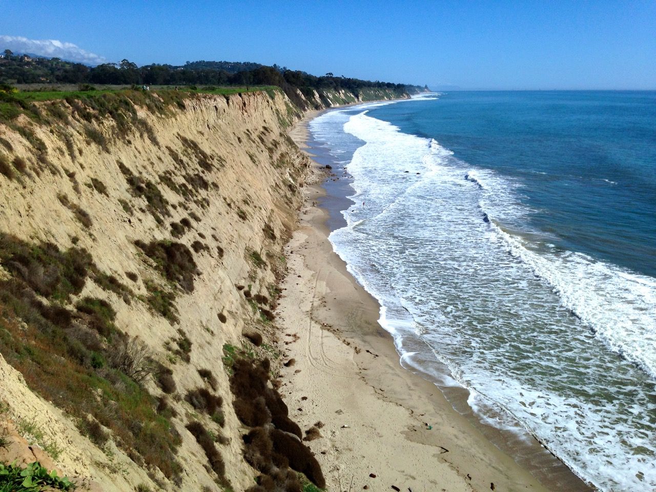 Sunny aerial view of steep sandy cliffs overlooking a narrow beach and ocean waves. - More Mesa Trail