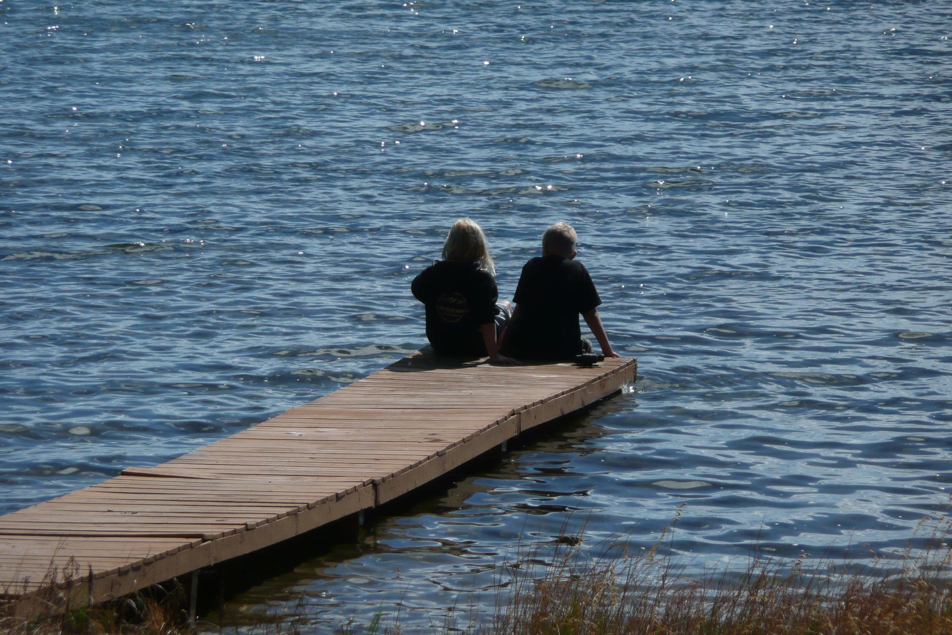 Two people sit on a wooden dock over calm blue water.