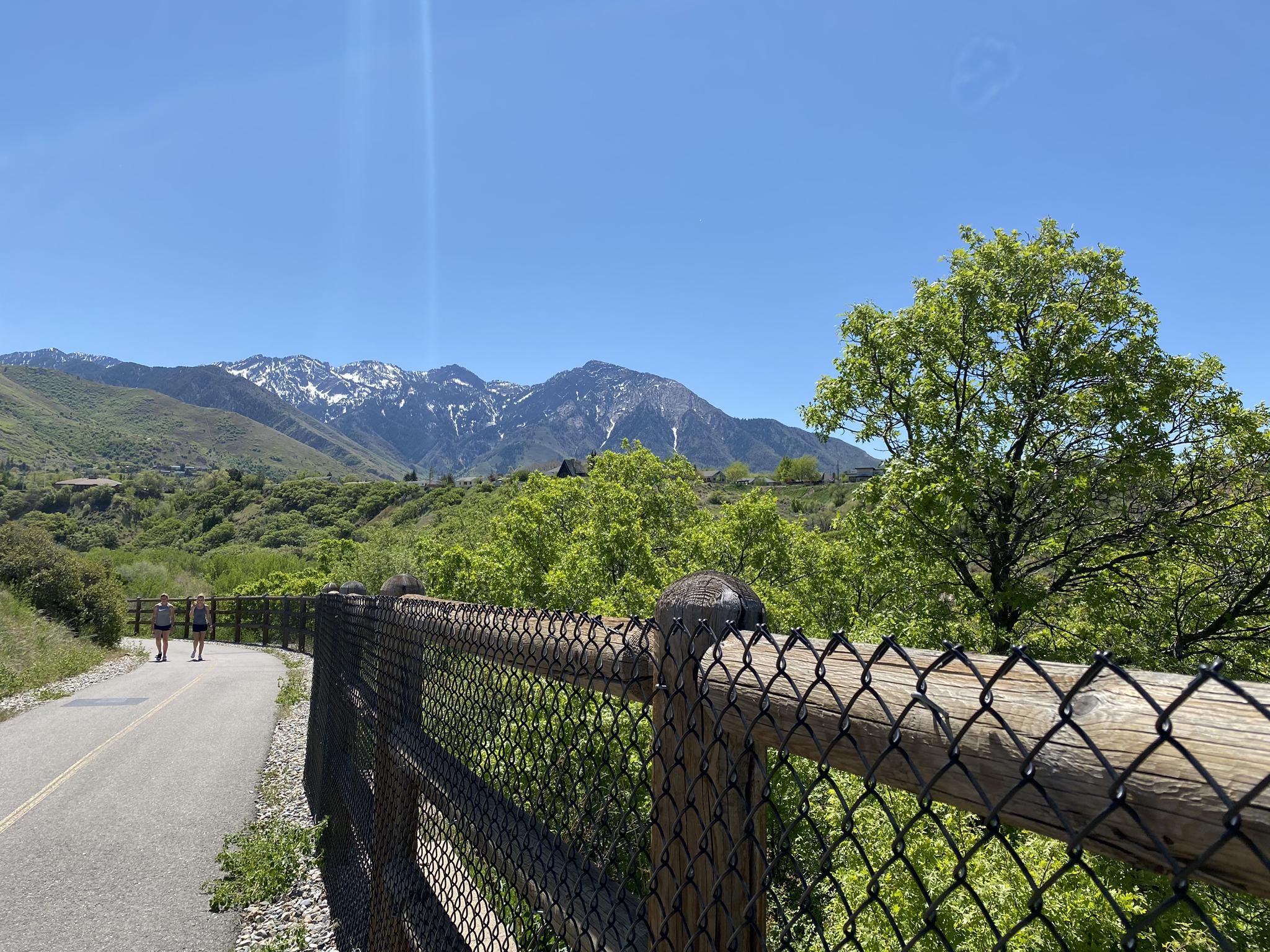 Two people walk on a sunny path next to a fence, with green mountains and trees. - Parley's Trail