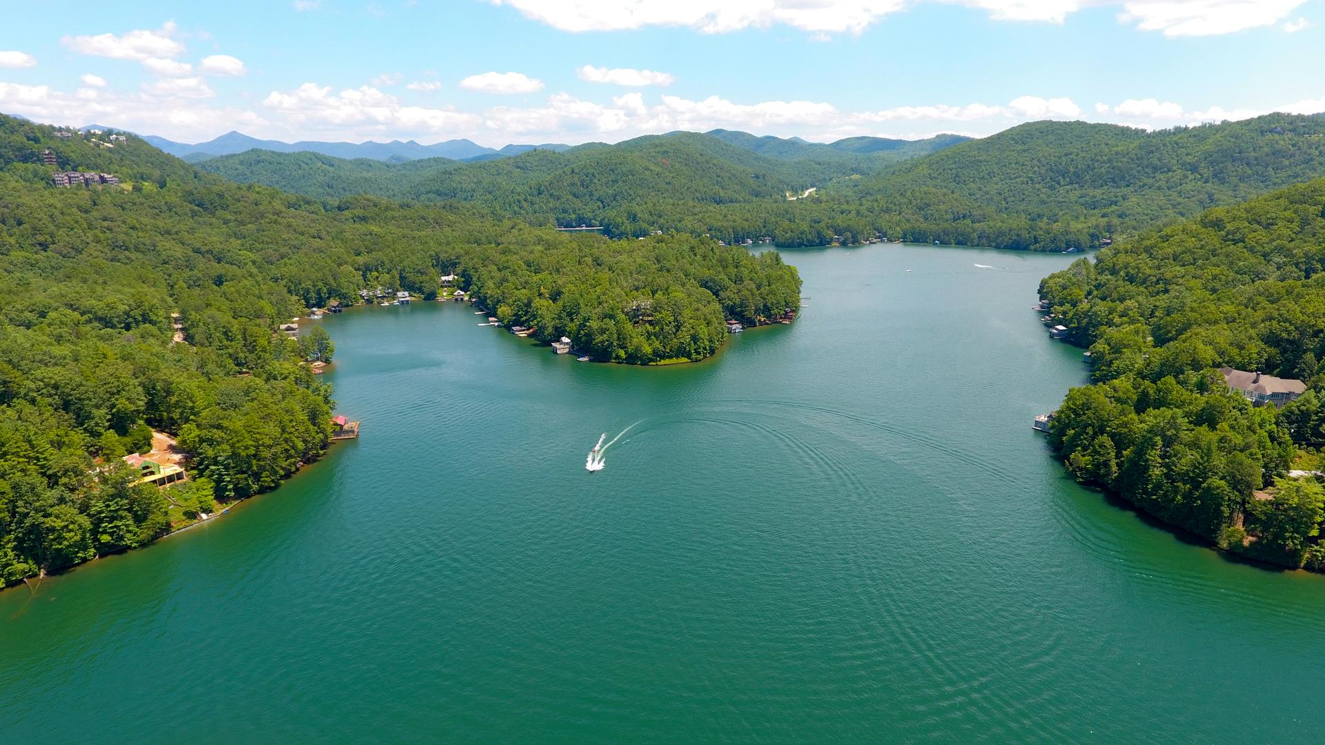 Aerial view of a boat on a sunny lake surrounded by green mountains.