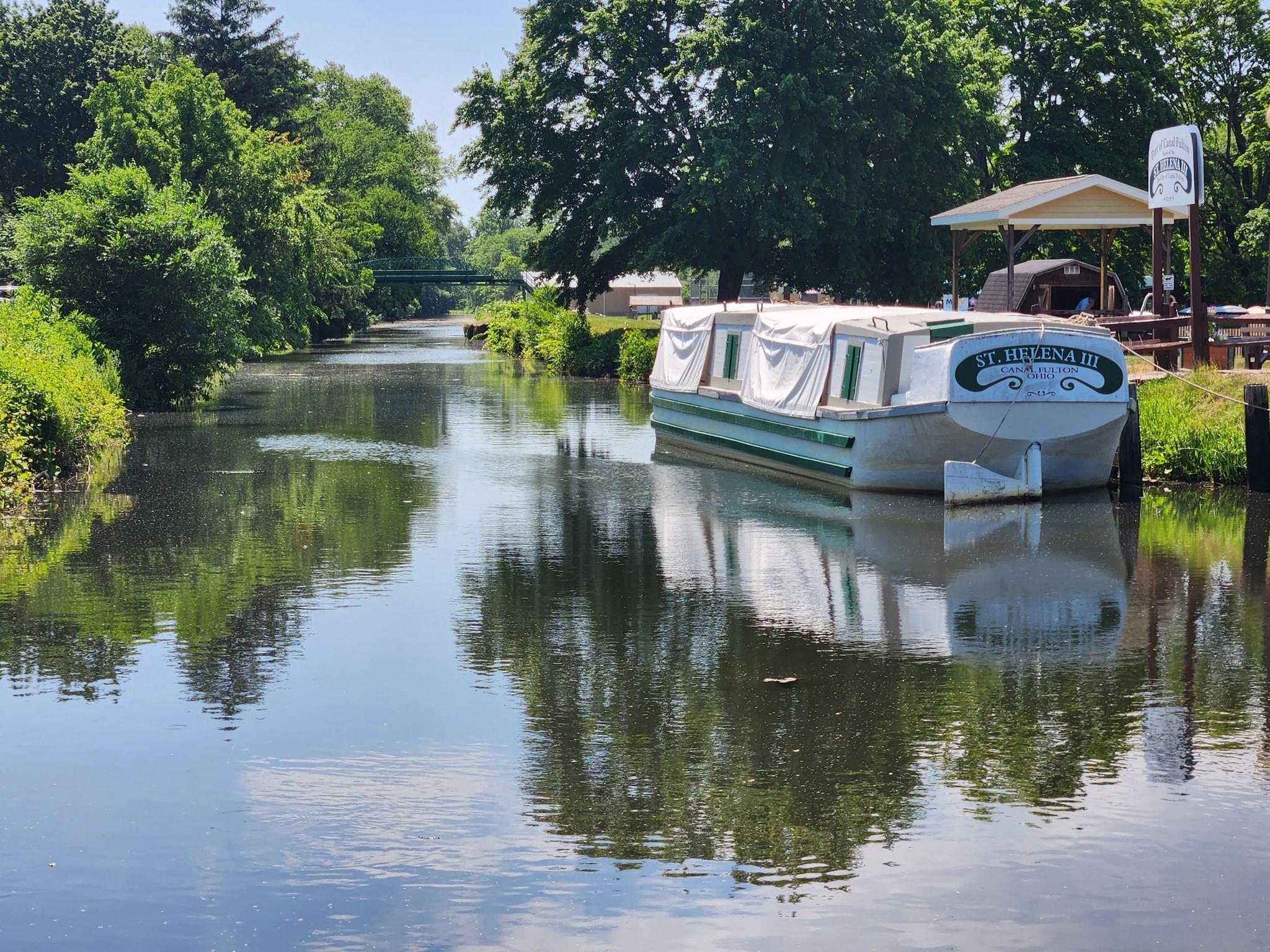 A white and green boat docked on a calm, tree-lined canal.
