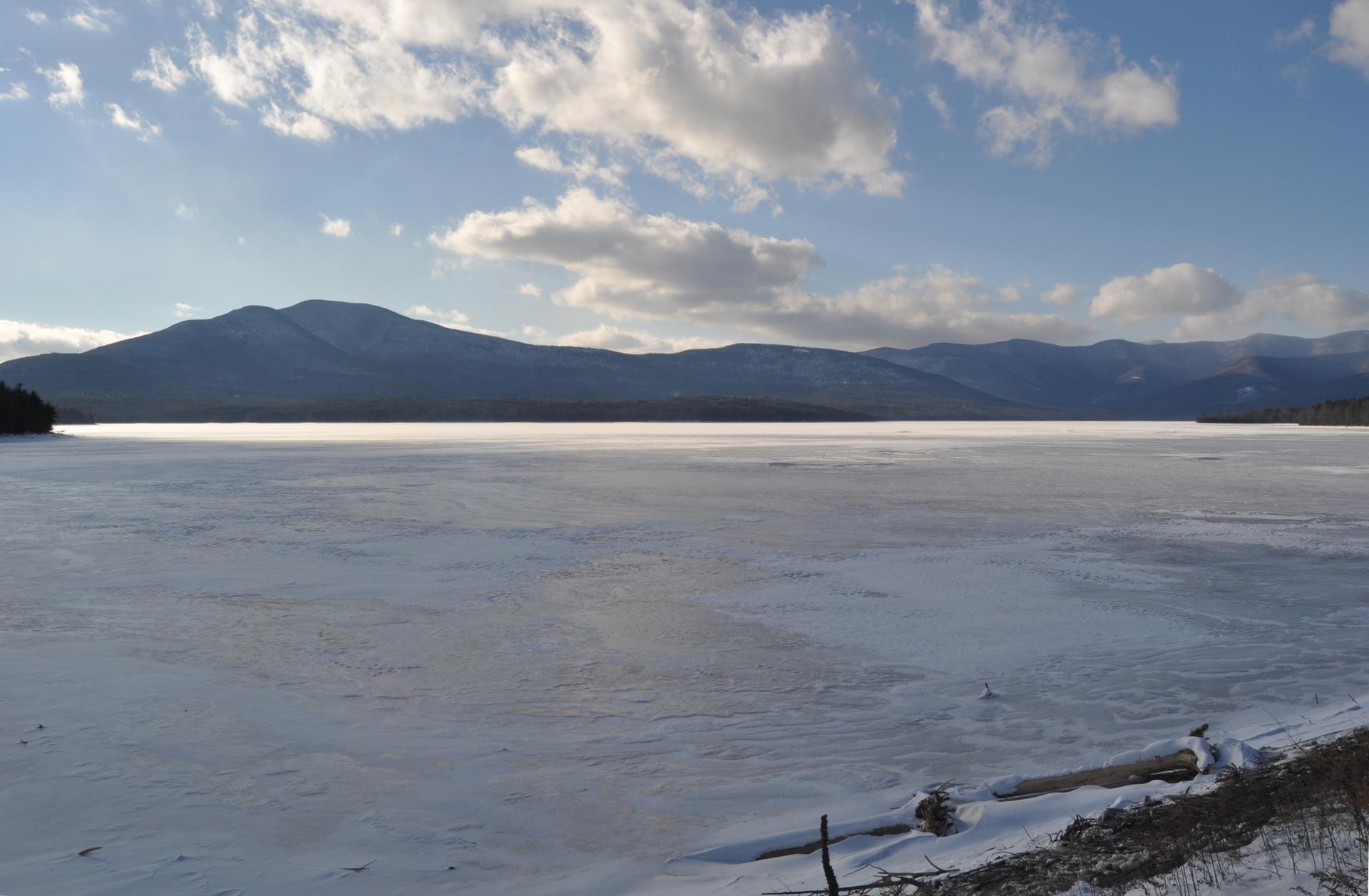 Expansive frozen lake, distant mountains, and a bright, cloudy winter sky.