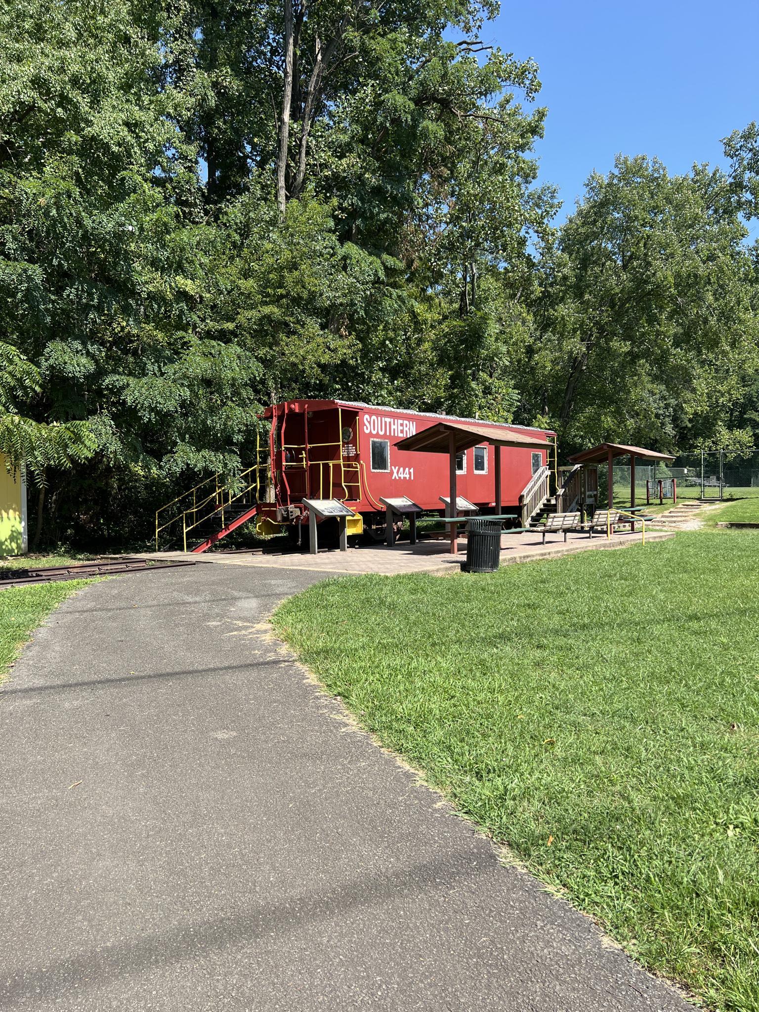 Red caboose beside a paved path, surrounded by green trees on a sunny day.