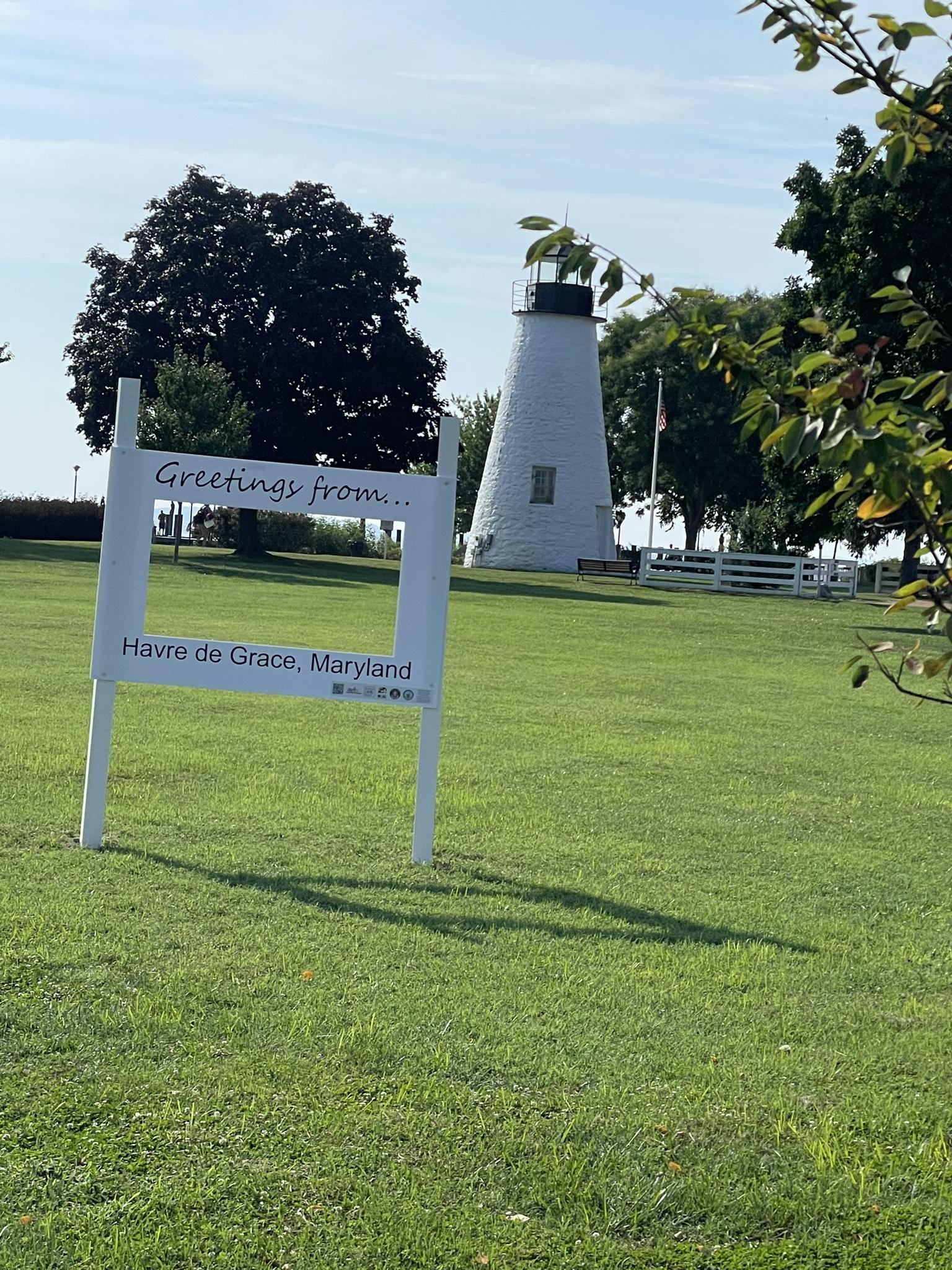 A white lighthouse stands in a sunny green field next to a welcome sign.