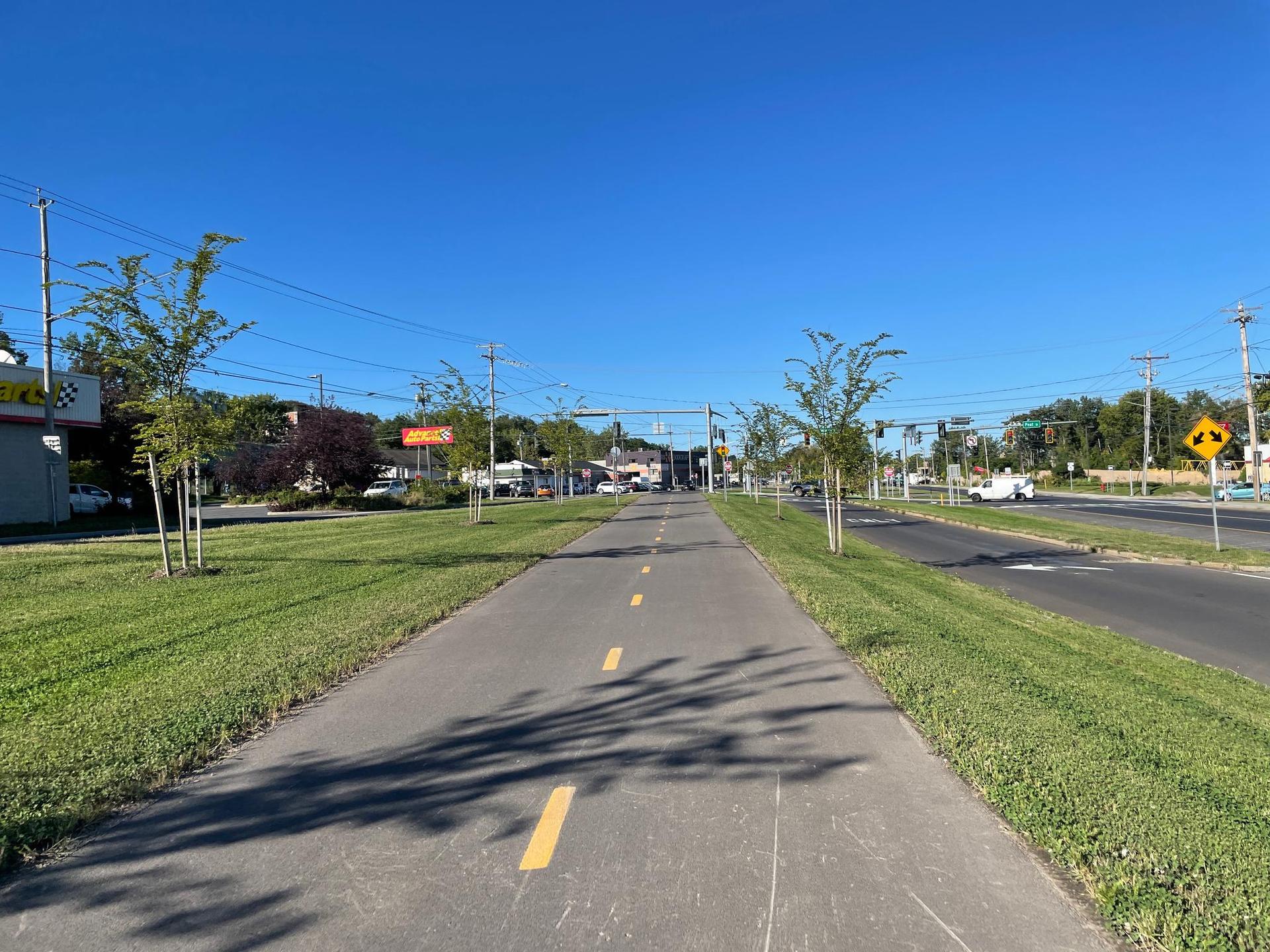 Paved path through a grassy area, with a road and buildings under a clear blue sky.