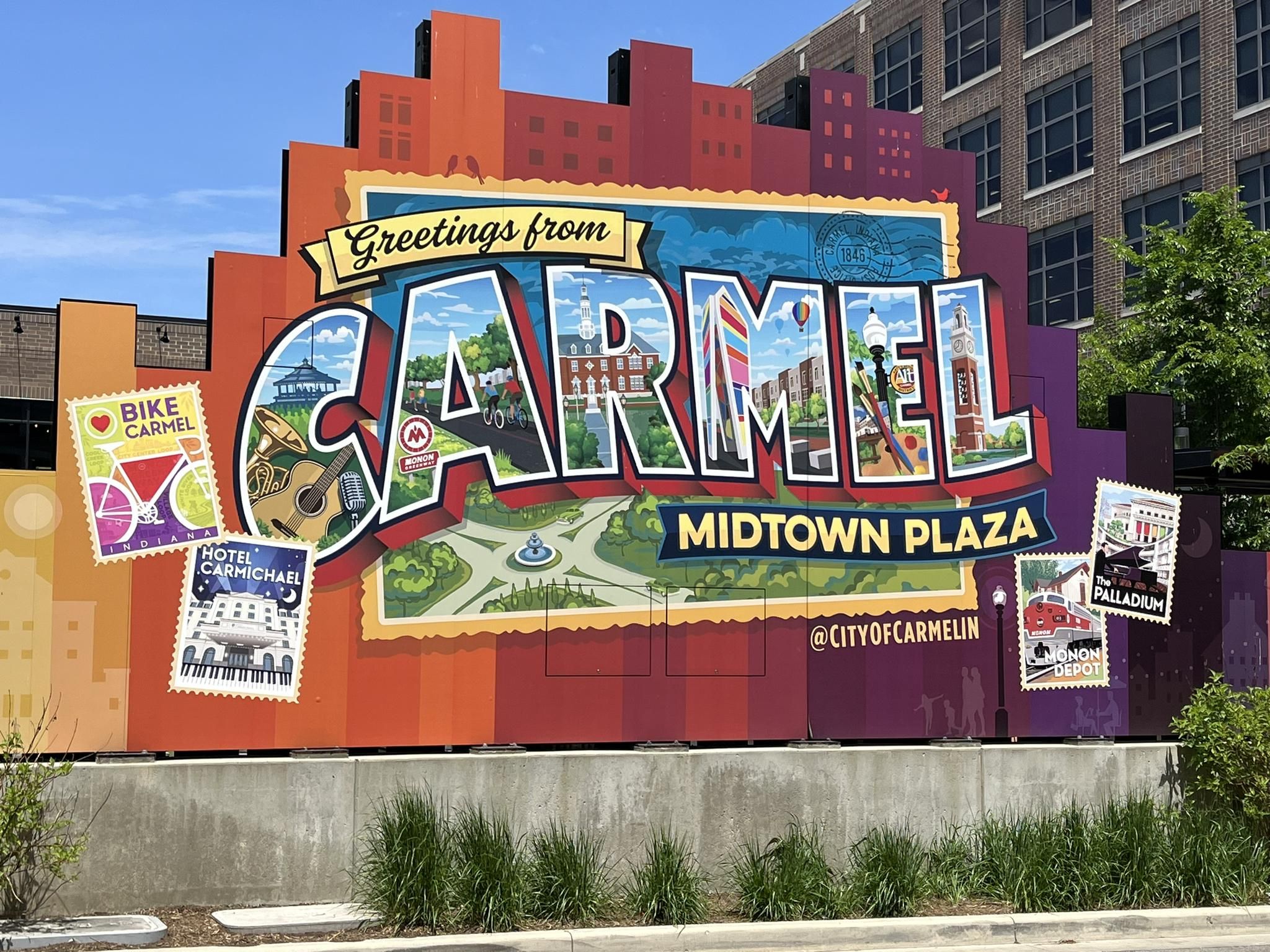 Large colorful postcard-style mural with "CARMEL" and local scenes on a sunny day. - Monon & Midland Trace Trails Loop
